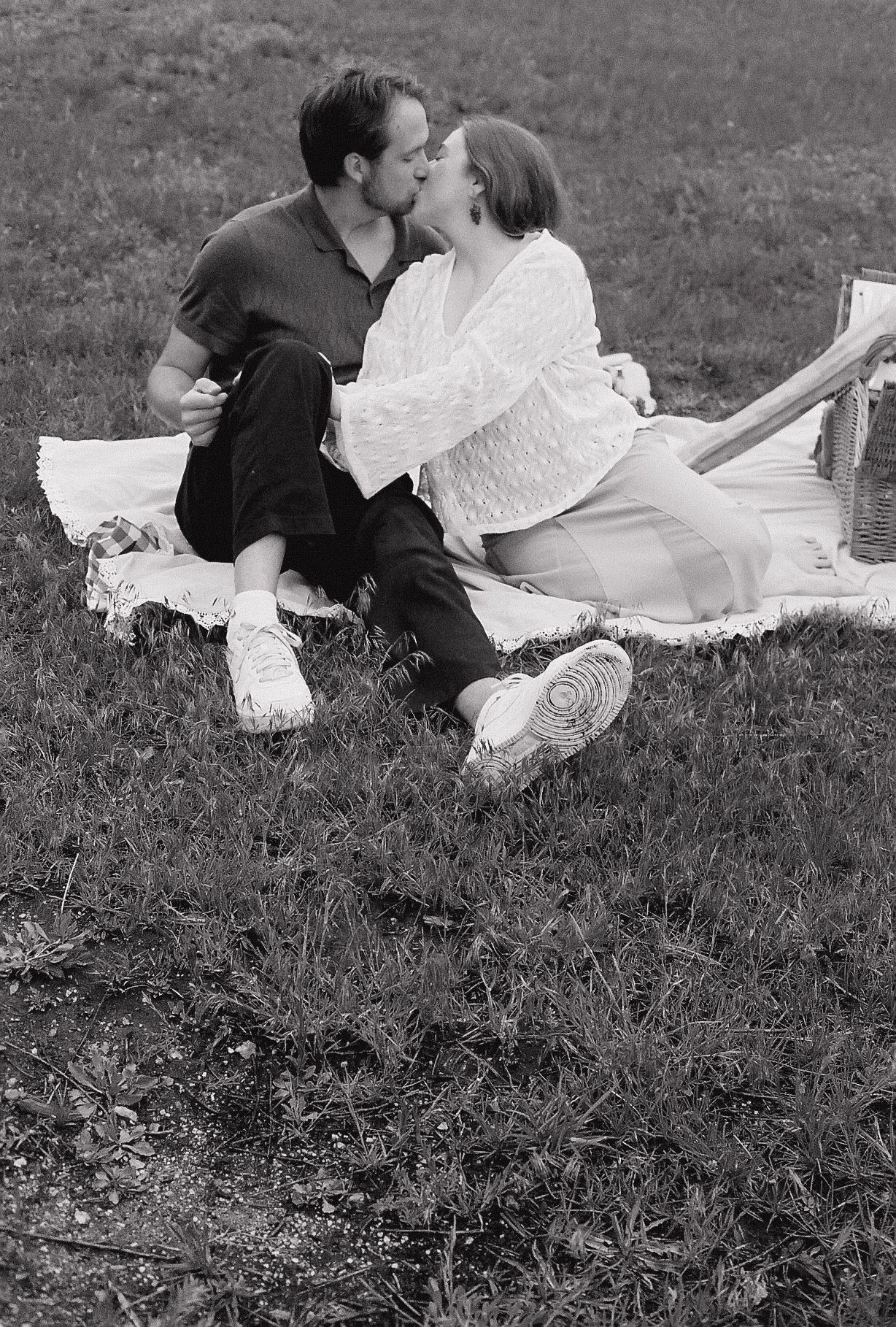 Black and white photo of couple sharing a kiss during a romantic outdoor picnic on grass.