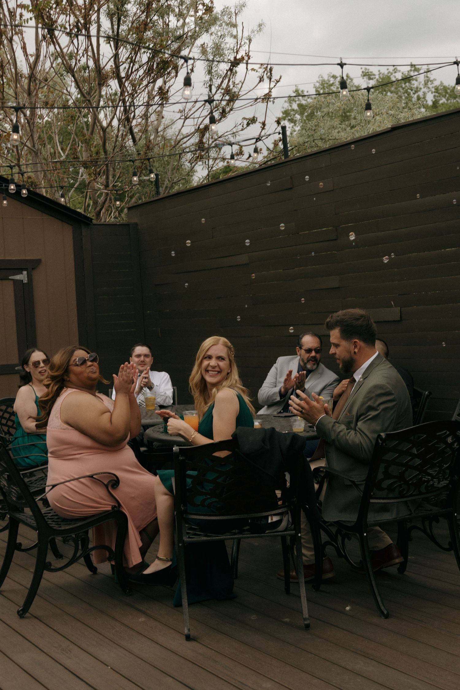 Wedding guests clap and celebrate together at outdoor reception tables on wooden deck with string lights.
