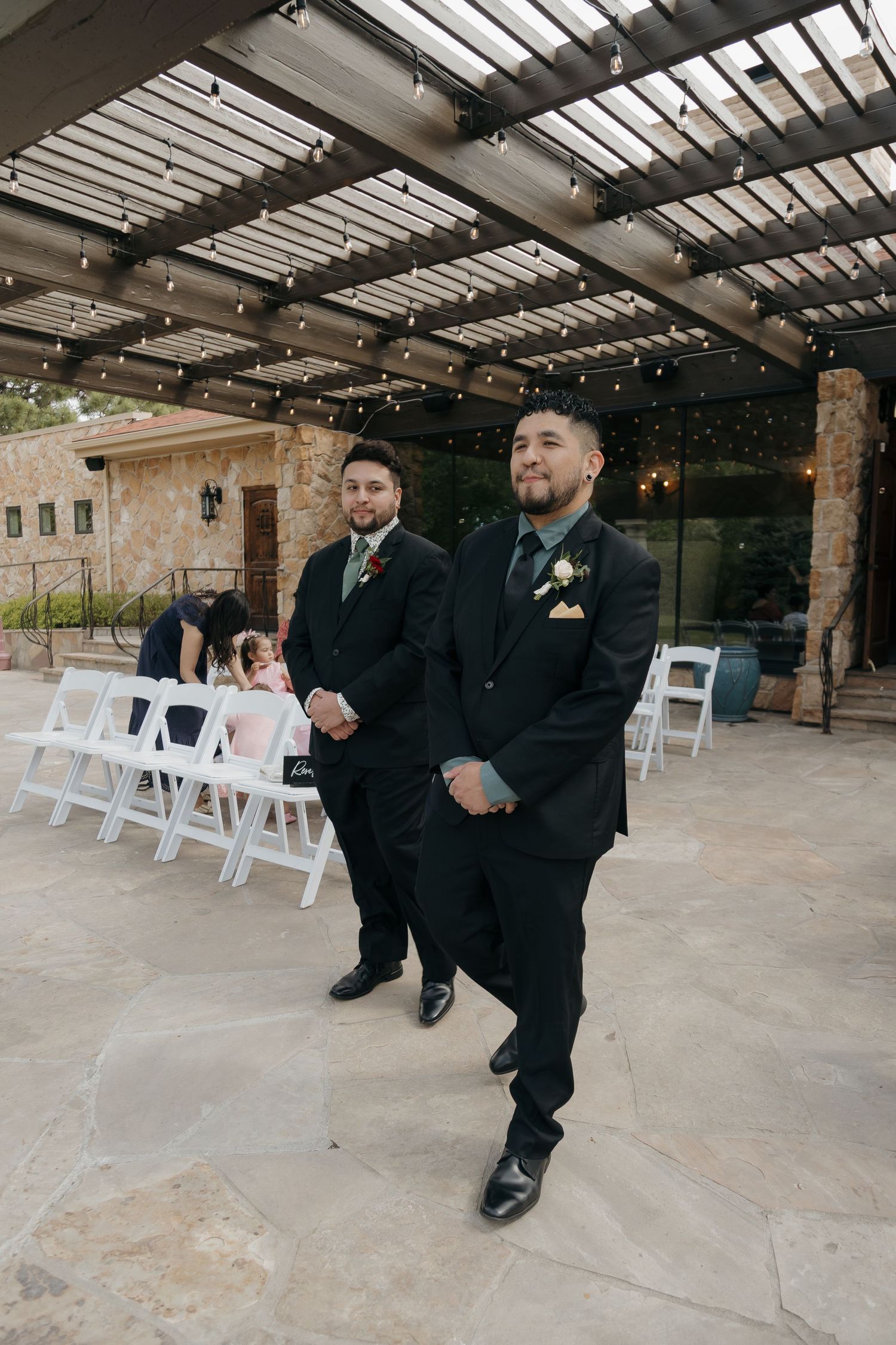 Groom stands confidently at outdoor wedding ceremony under a pergola with string lights.