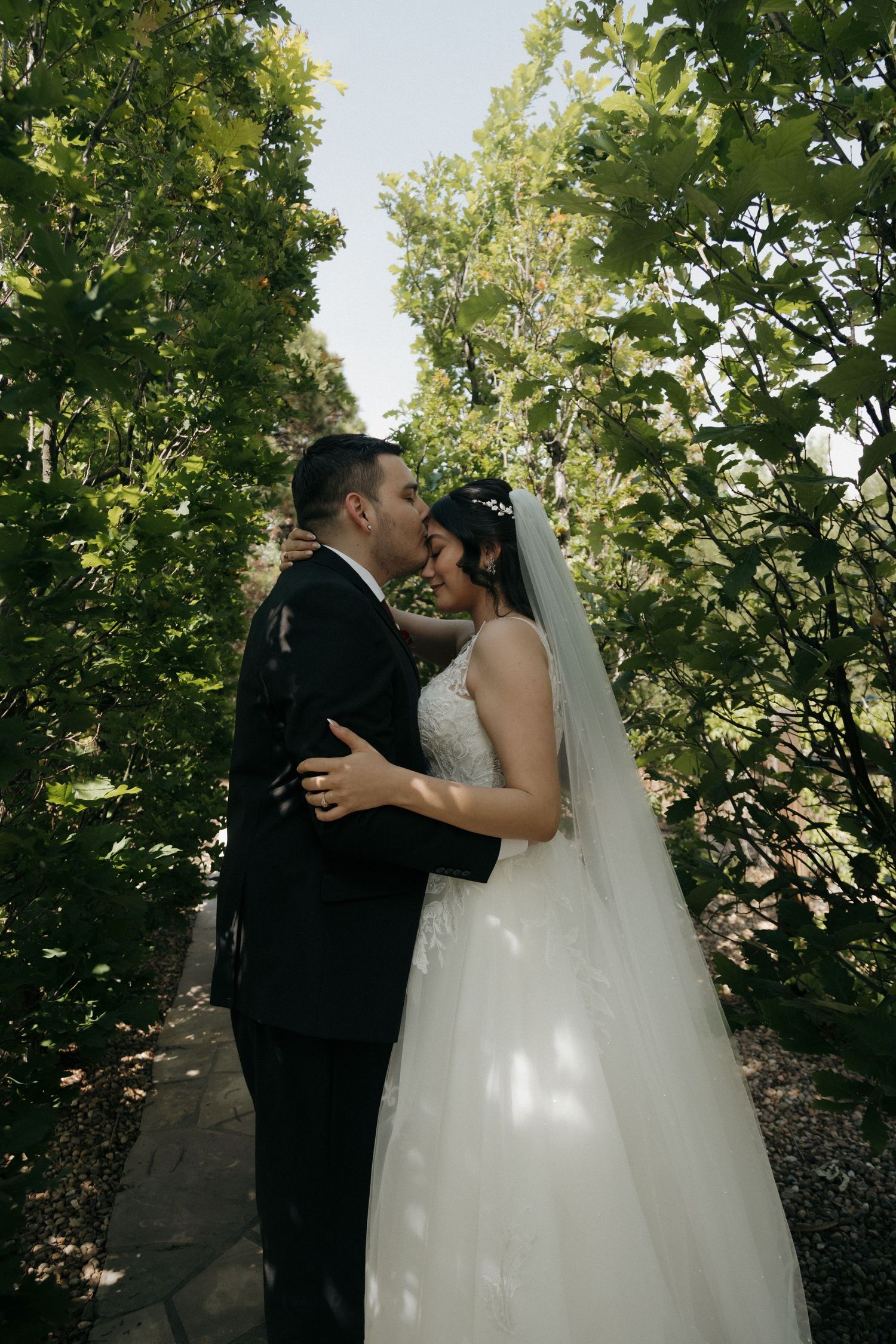 Bride and groom sharing a romantic kiss on a sunlit garden pathway surrounded by lush greenery.