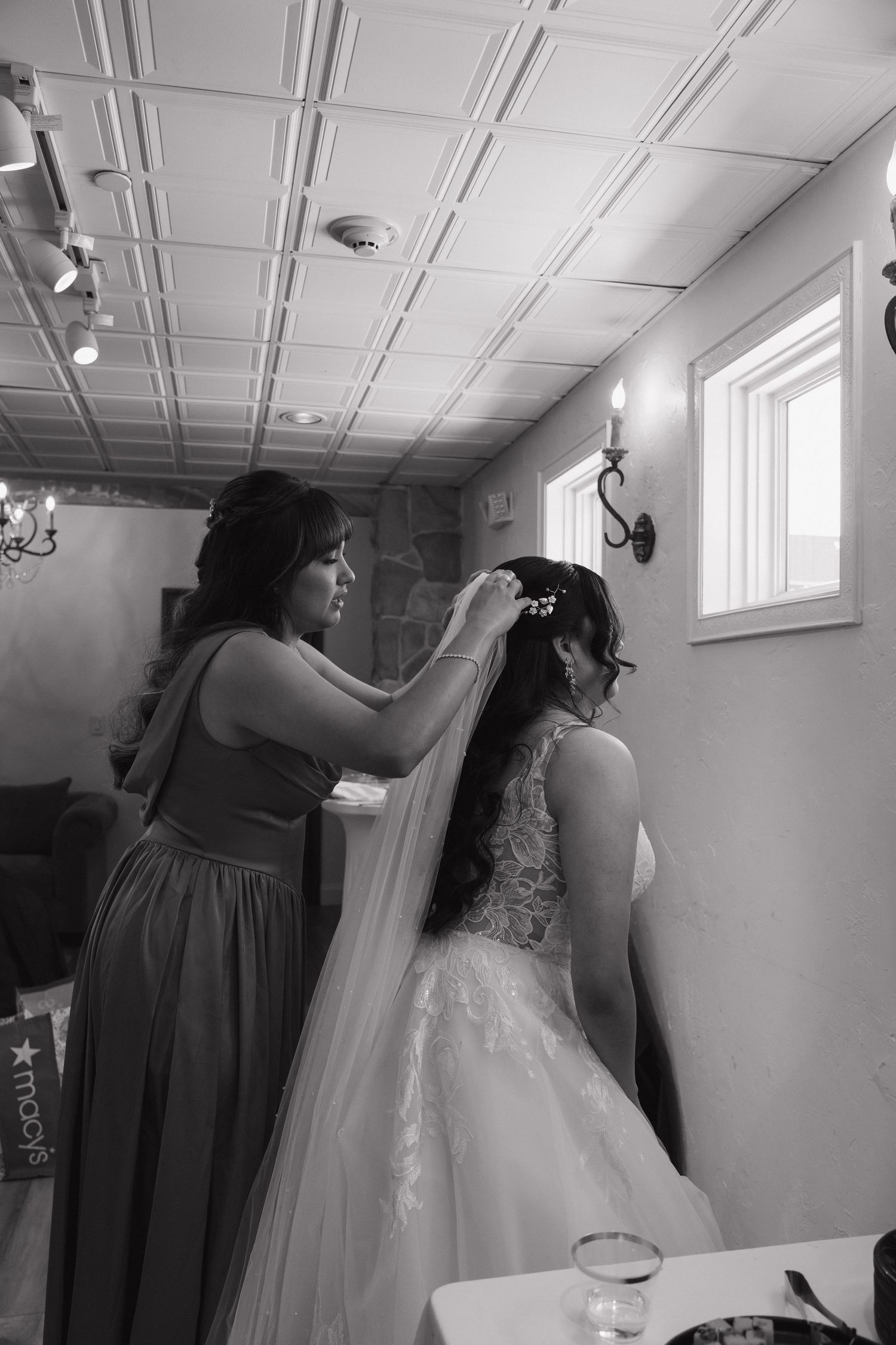 Bridesmaid adjusts the veil of a bride in a lace wedding gown in a black and white wedding preparation photo.