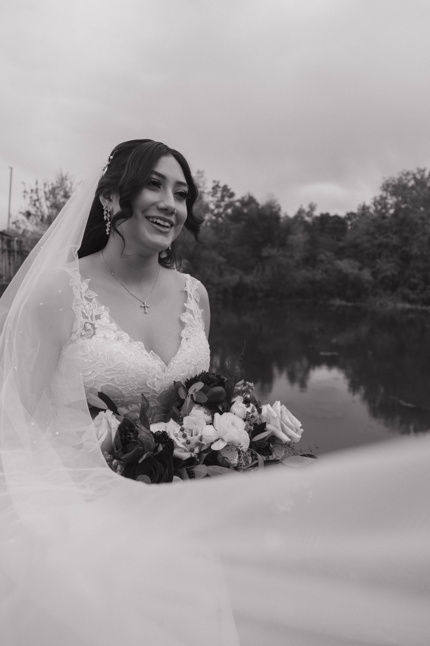 Black and white portrait of smiling bride holding a floral bouquet with flowing veil by a reflective lake.