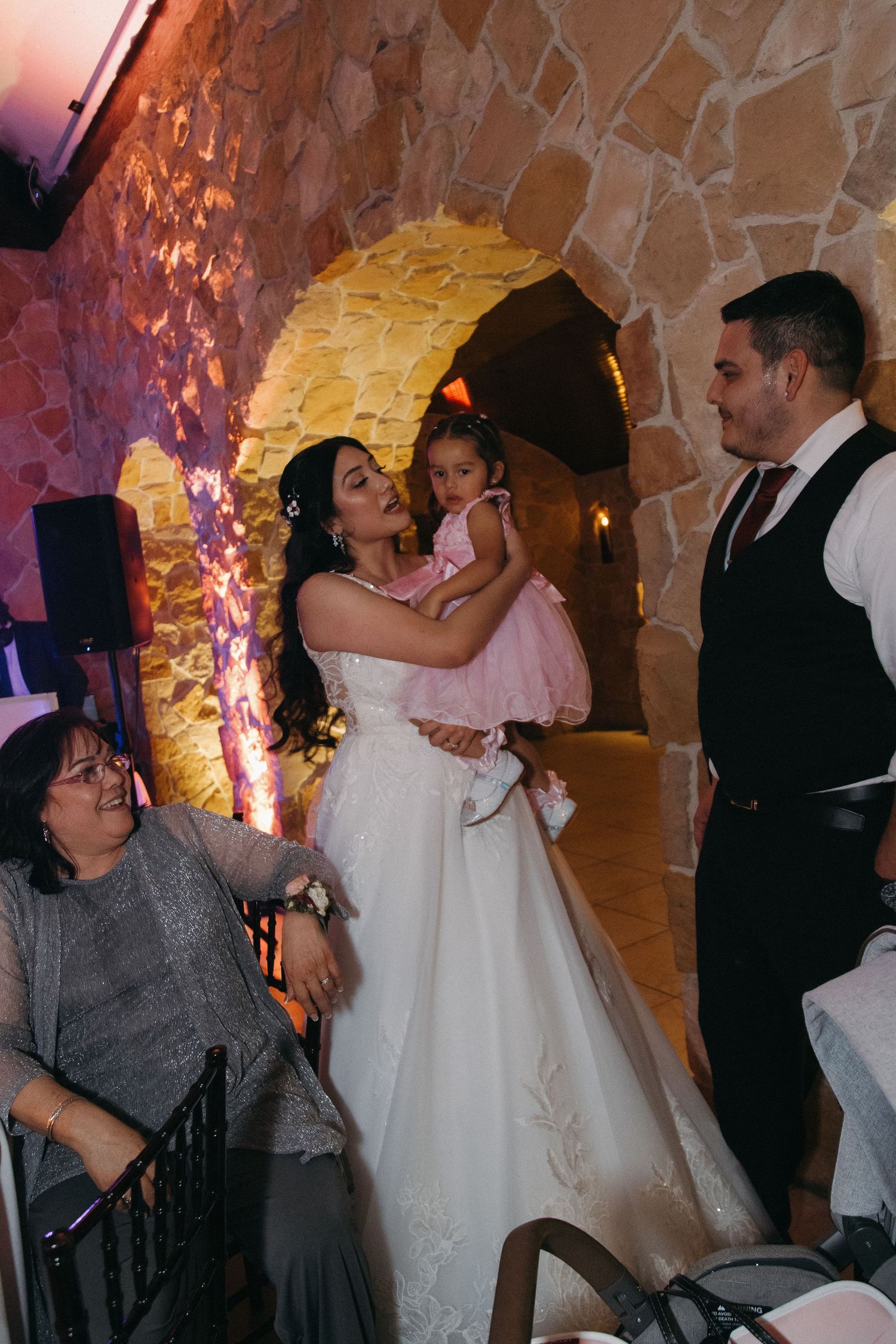 Bride in lace gown holds young girl in pink dress while groom stands nearby in stone-walled reception hall.