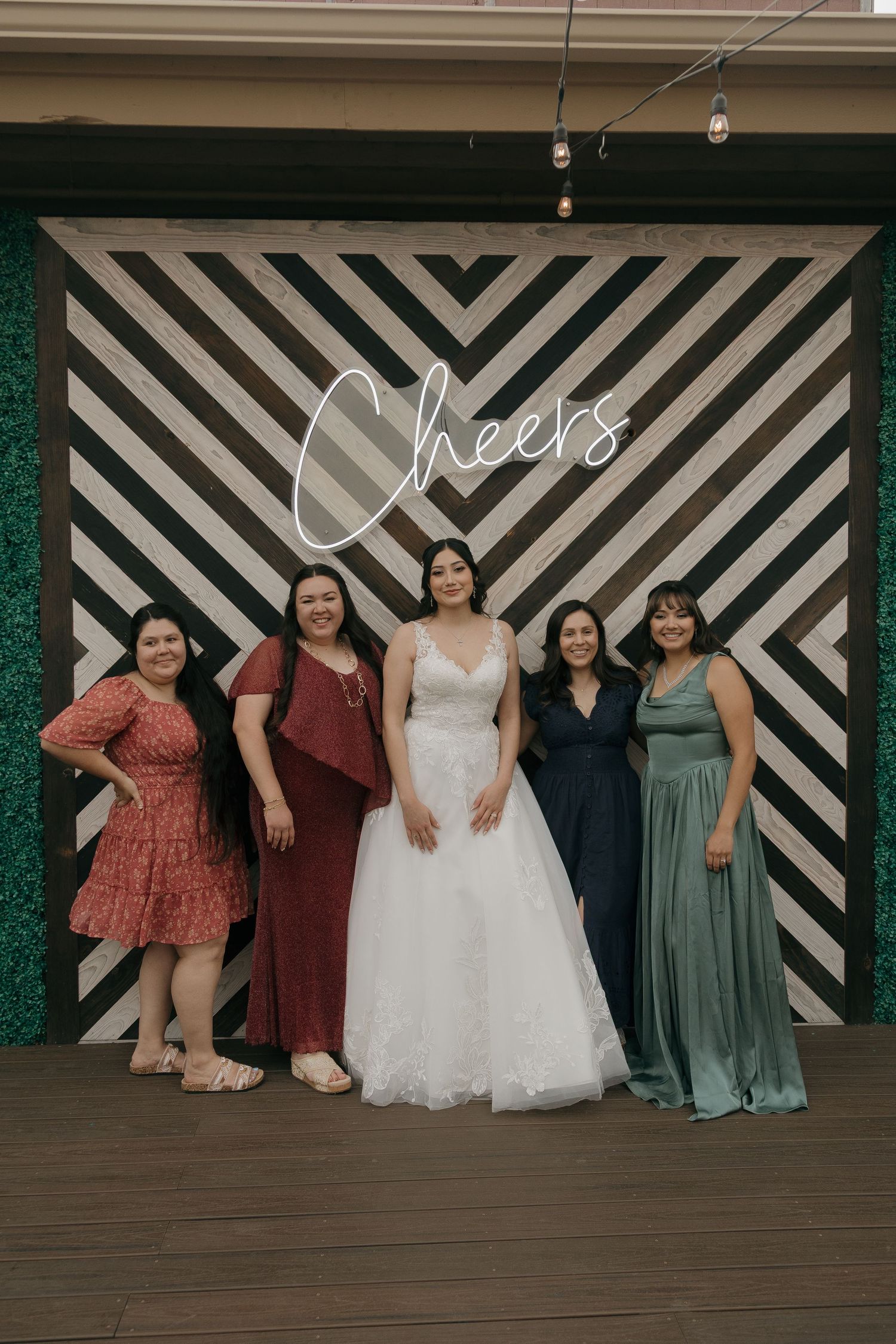 Bride in white ball gown poses with four guests in front of a Cheers neon sign at wedding reception.
