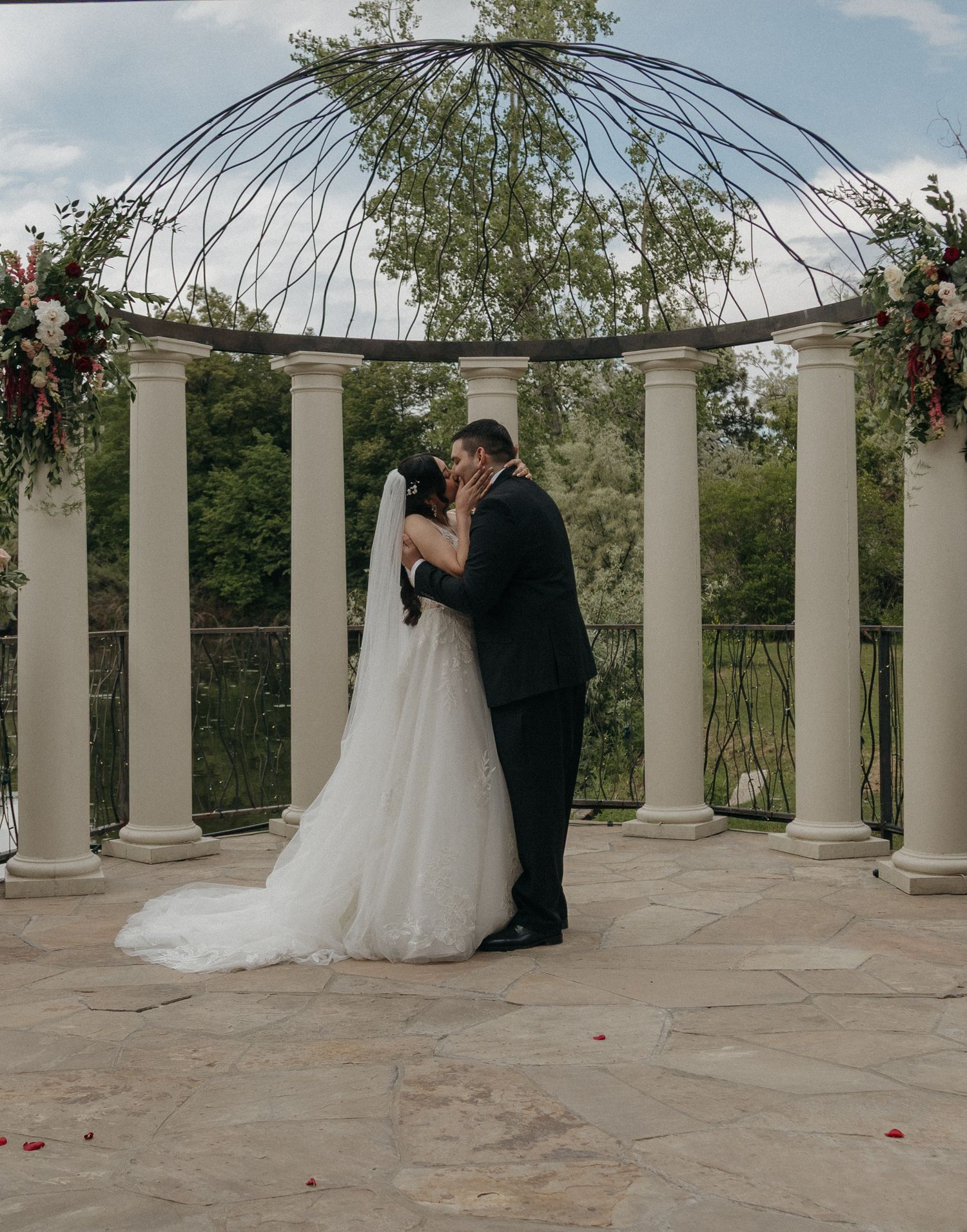 Newlyweds share first kiss under ornate iron and column gazebo at outdoor wedding ceremony.