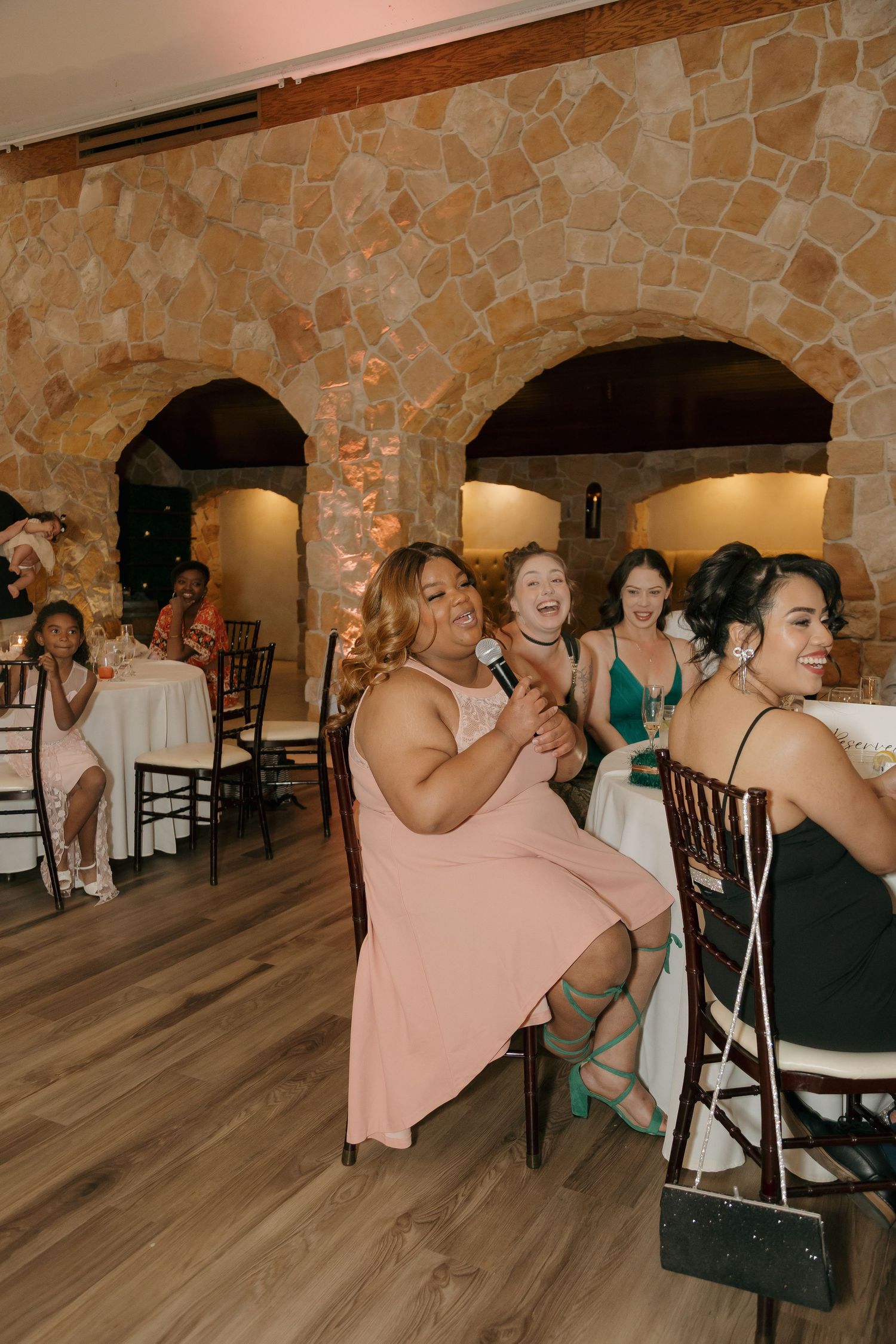 Wedding guest in pink dress sings into microphone laughing at rustic stone venue reception.