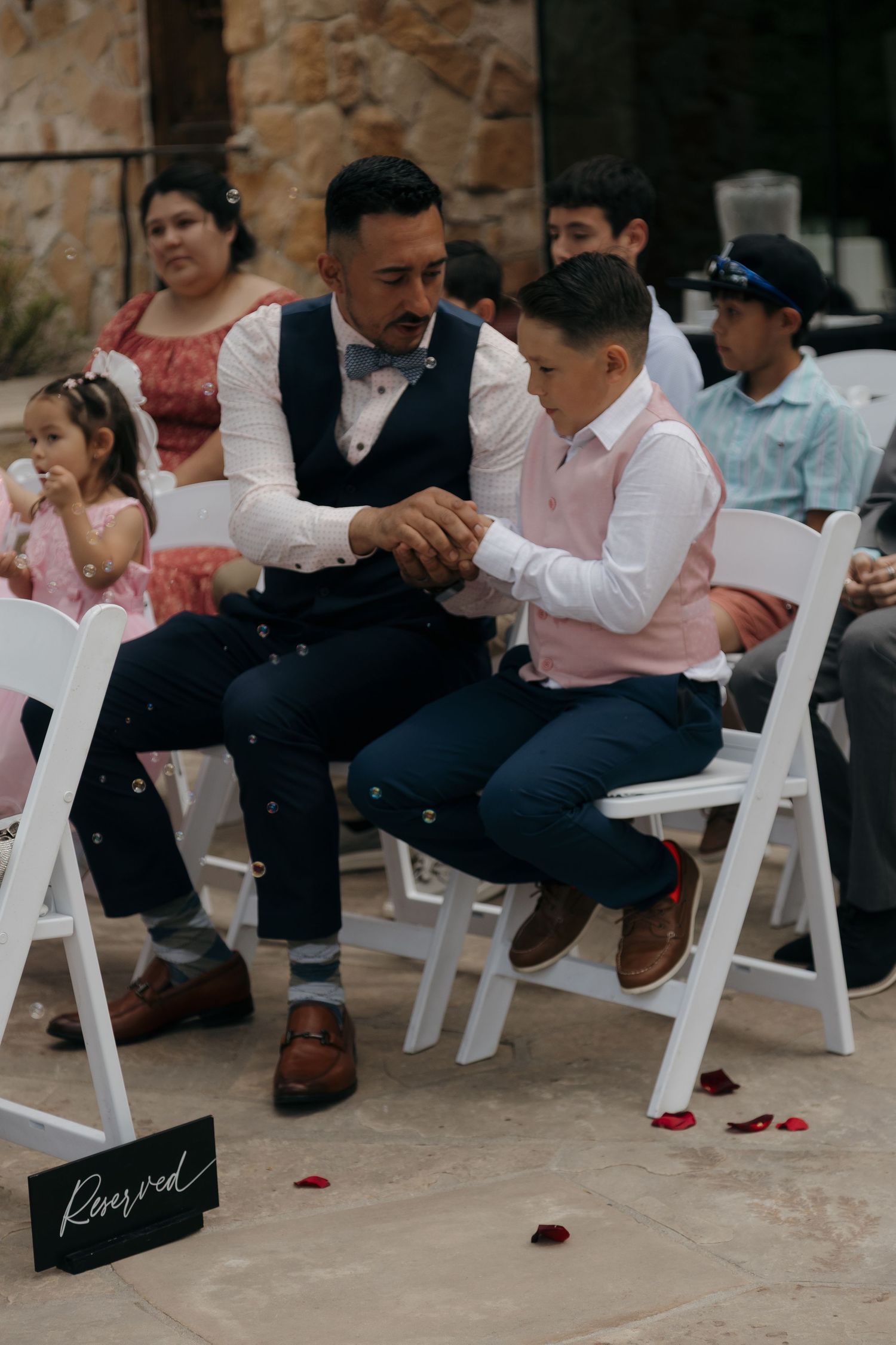 Father and son in formal attire sitting in reserved seats at outdoor wedding ceremony.