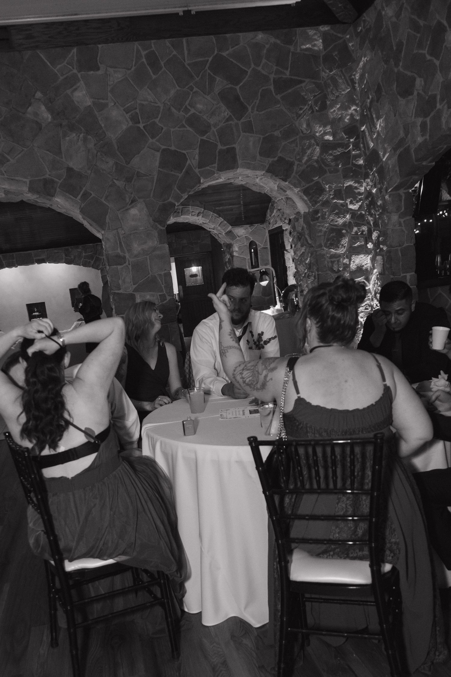 Wedding guests gather around table in rustic stone arched venue during black and white reception photo.