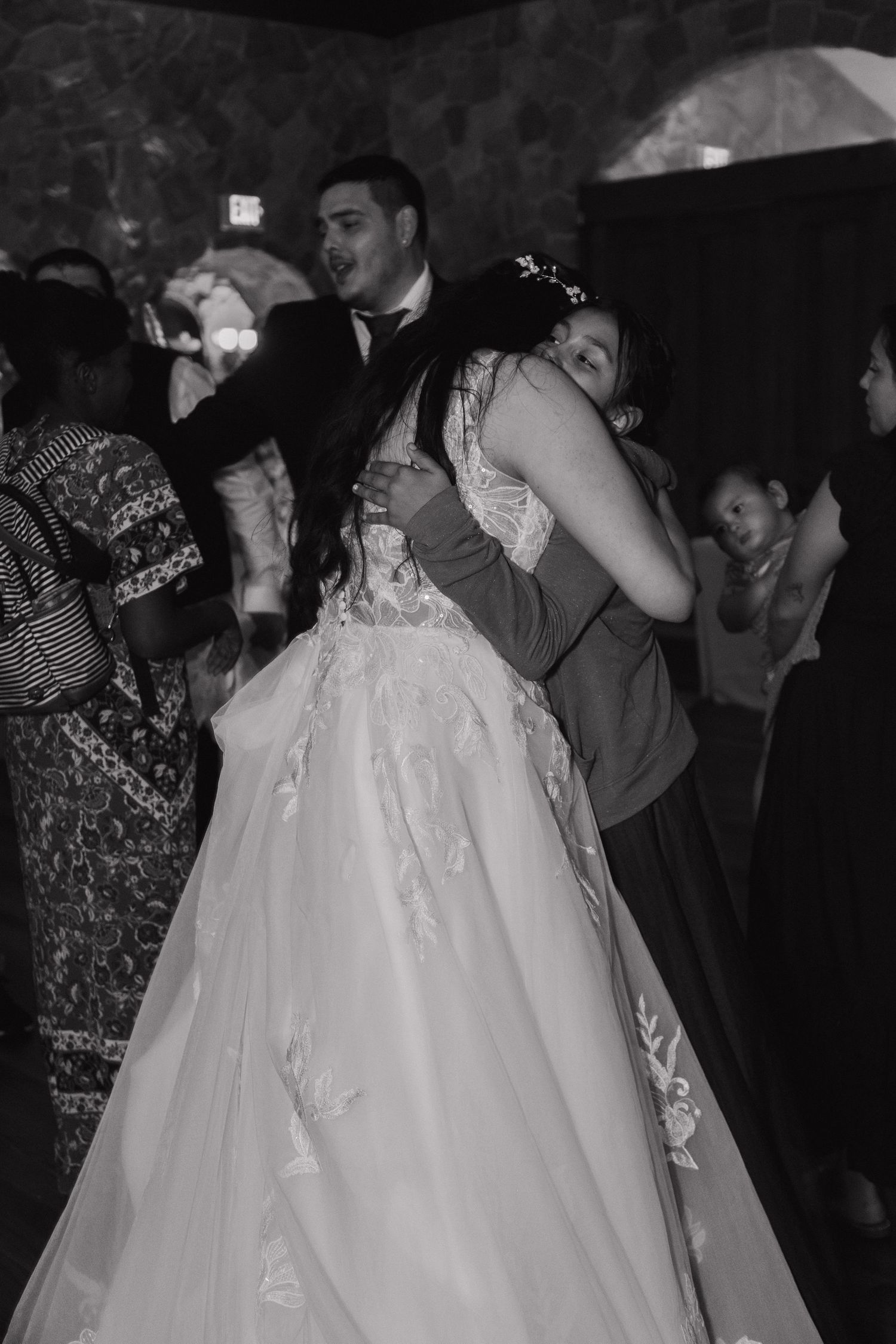 Black and white photo of bride and groom embracing on crowded wedding reception dance floor.