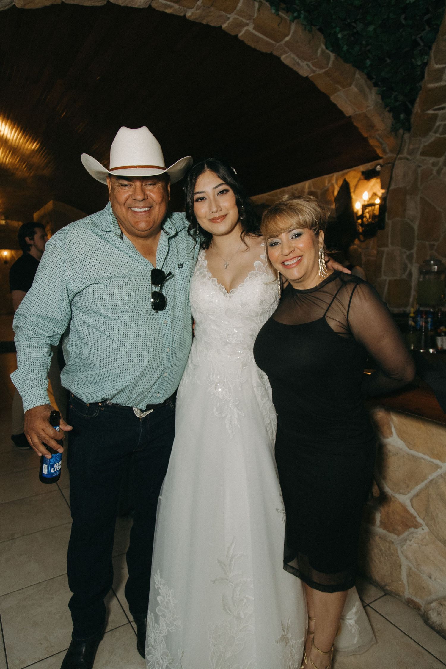 Bride in white lace gown poses with two guests, one wearing a cowboy hat, at a wedding reception.