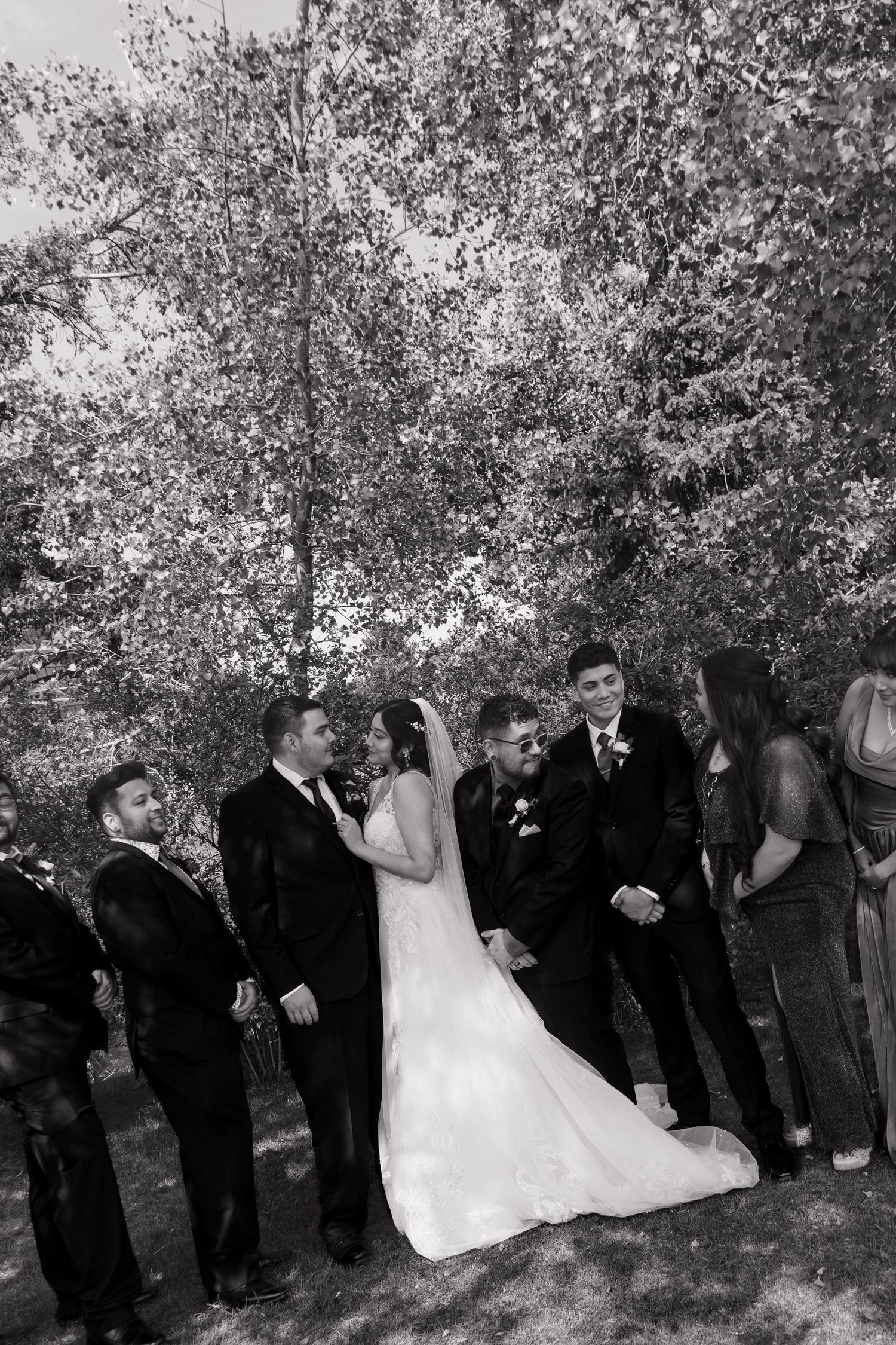 Black and white photo of bride, groom, and wedding party laughing together outdoors beneath tall trees.