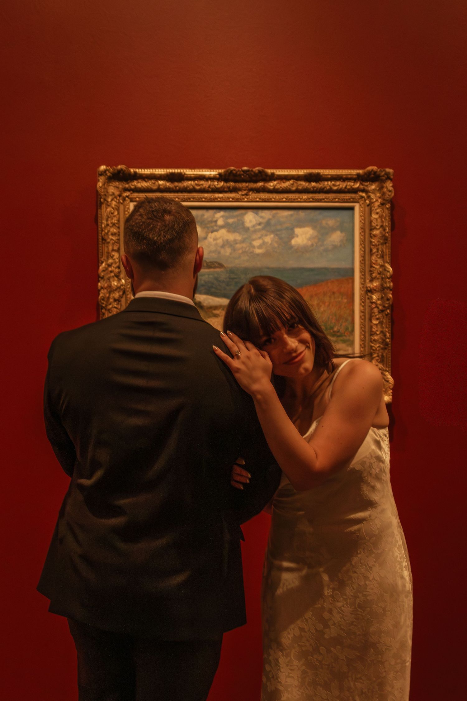 Two people in formal attire pose in front of golden framed paintings on a red wall.