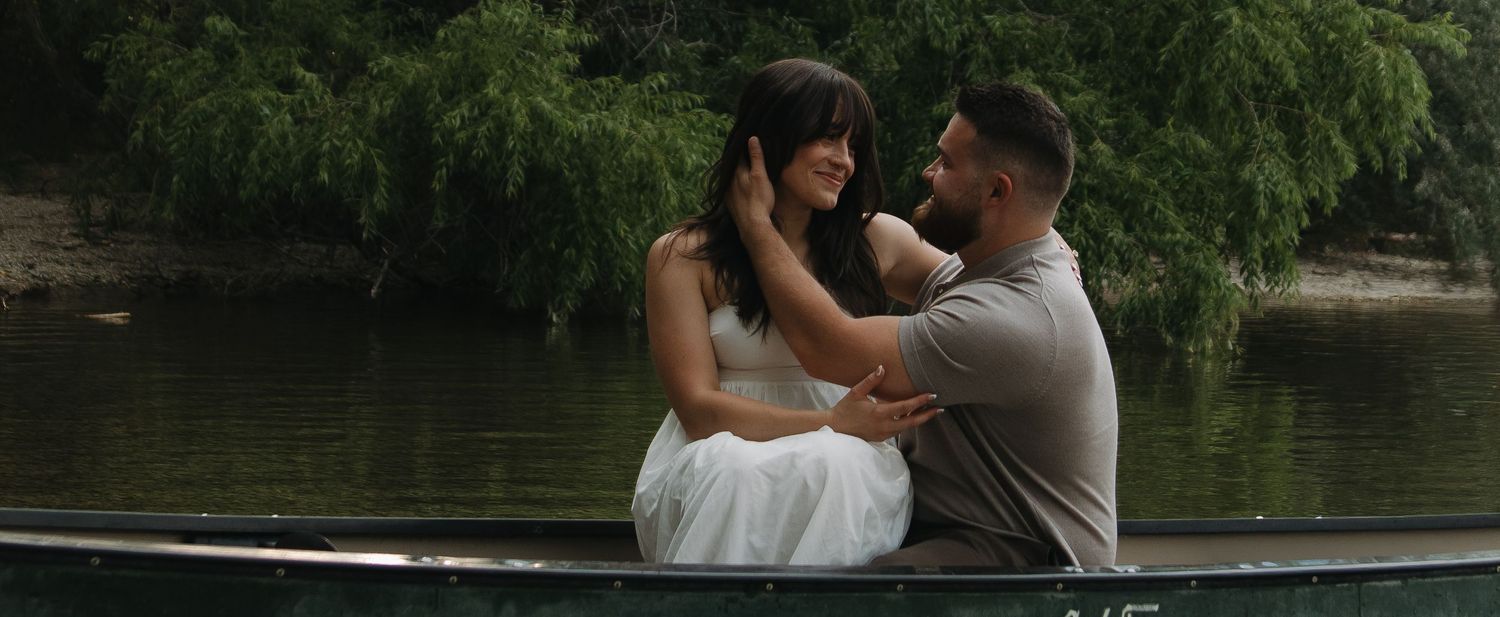 A couple stands in a white boat on a tranquil lake surrounded by lush greenery and trees.