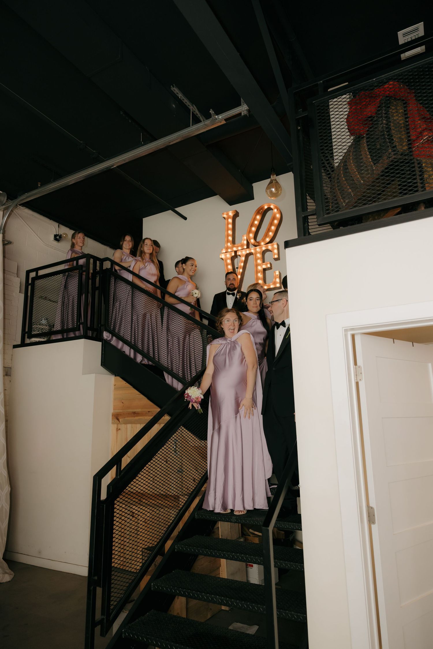 Wedding party descending industrial staircase with illuminated LOVE marquee sign in background.