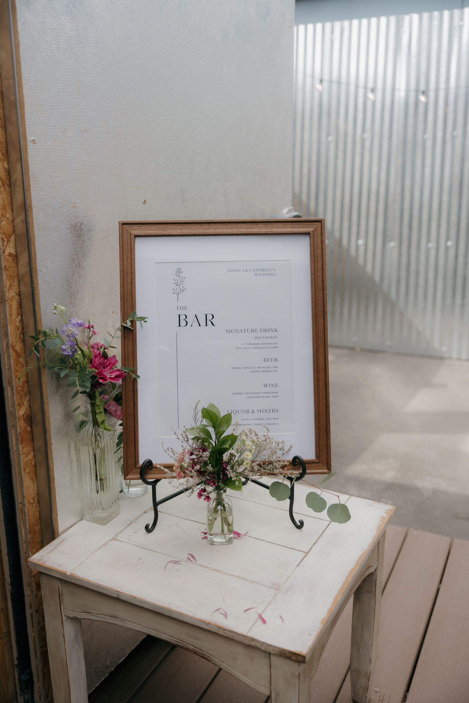 Elegant wedding bar menu sign in wooden frame displayed on white table with fresh floral arrangements.