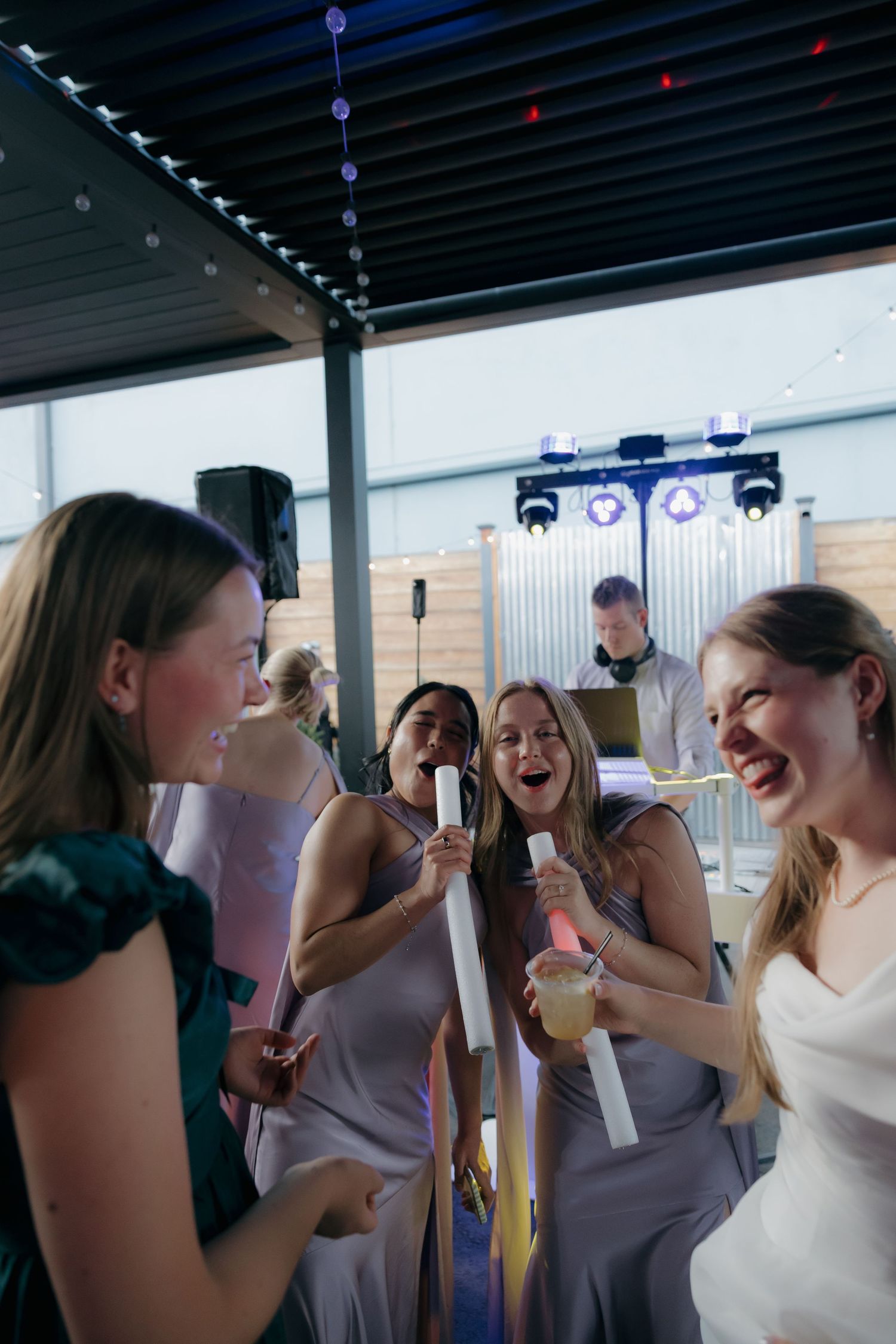Bridesmaids in lavender dresses laughing and singing with microphones alongside bride at wedding reception.