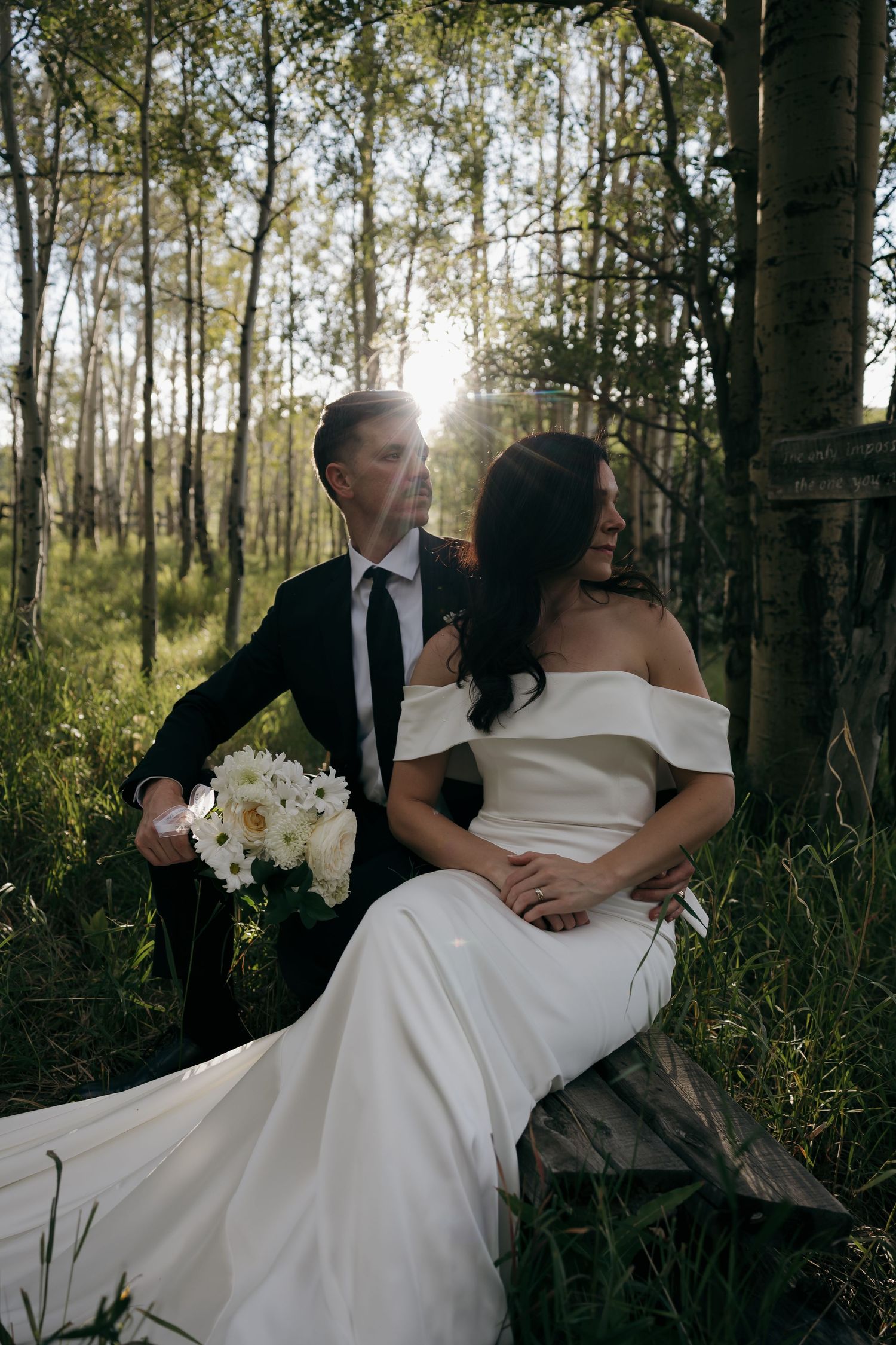 Bride in elegant off-shoulder gown sits with groom in black suit among sunlit aspen trees.