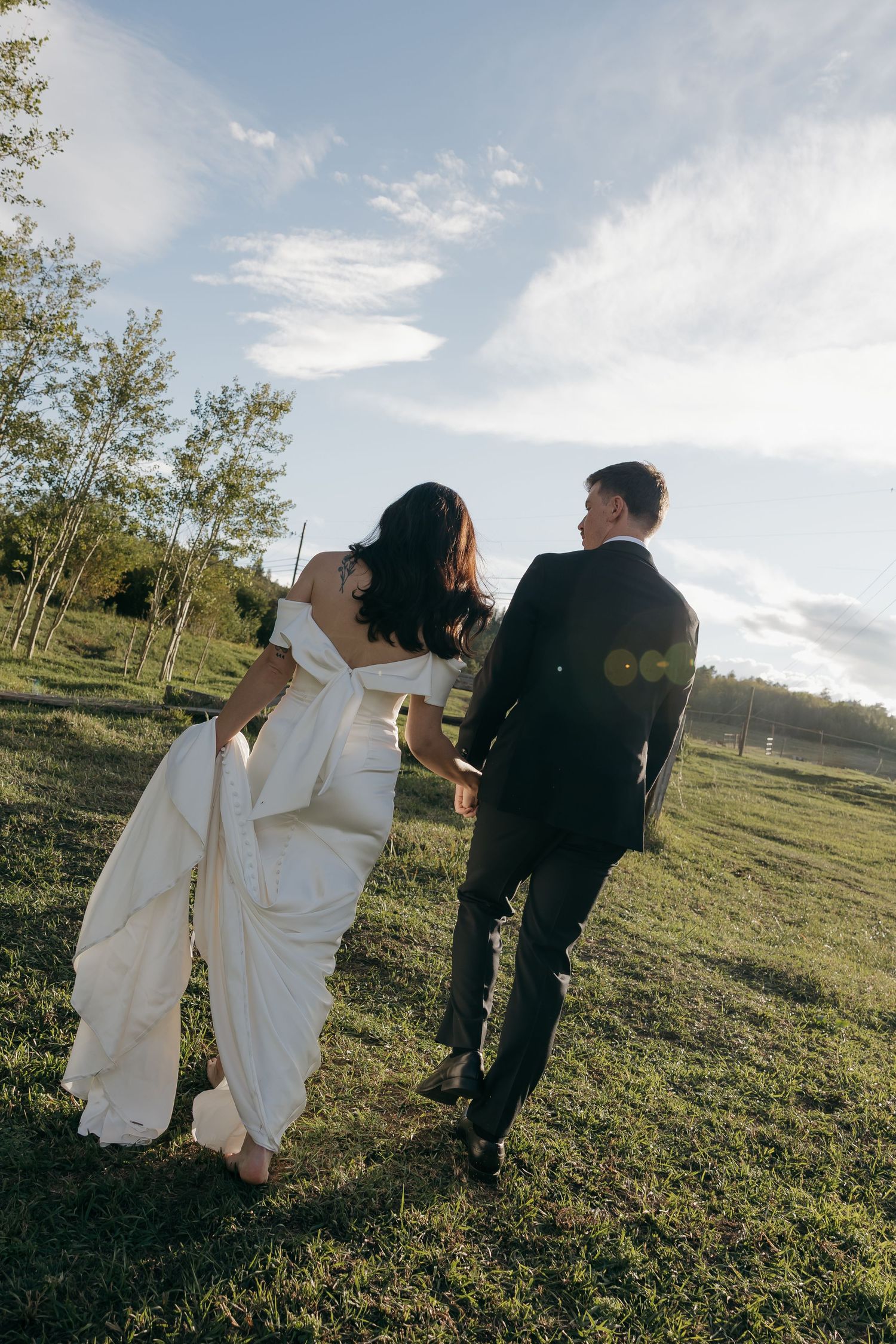 Bride and groom walk hand-in-hand across lush green field at sunset, bride lifting white gown.