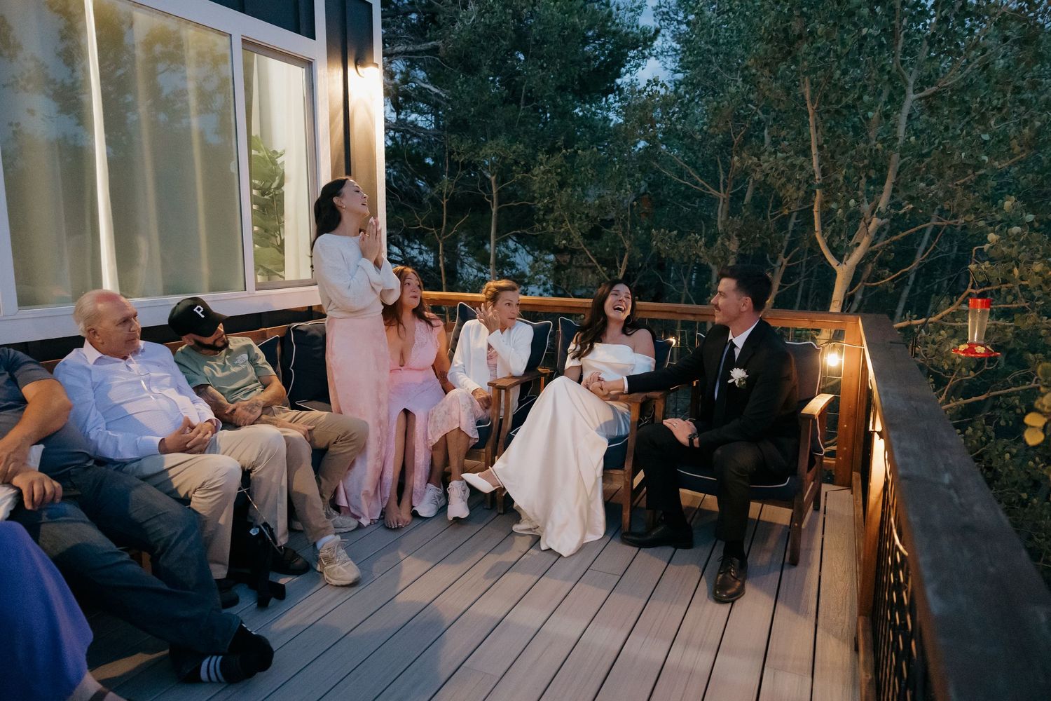 Wedding couple seated on outdoor deck with guests during evening celebration surrounded by trees and string lights.