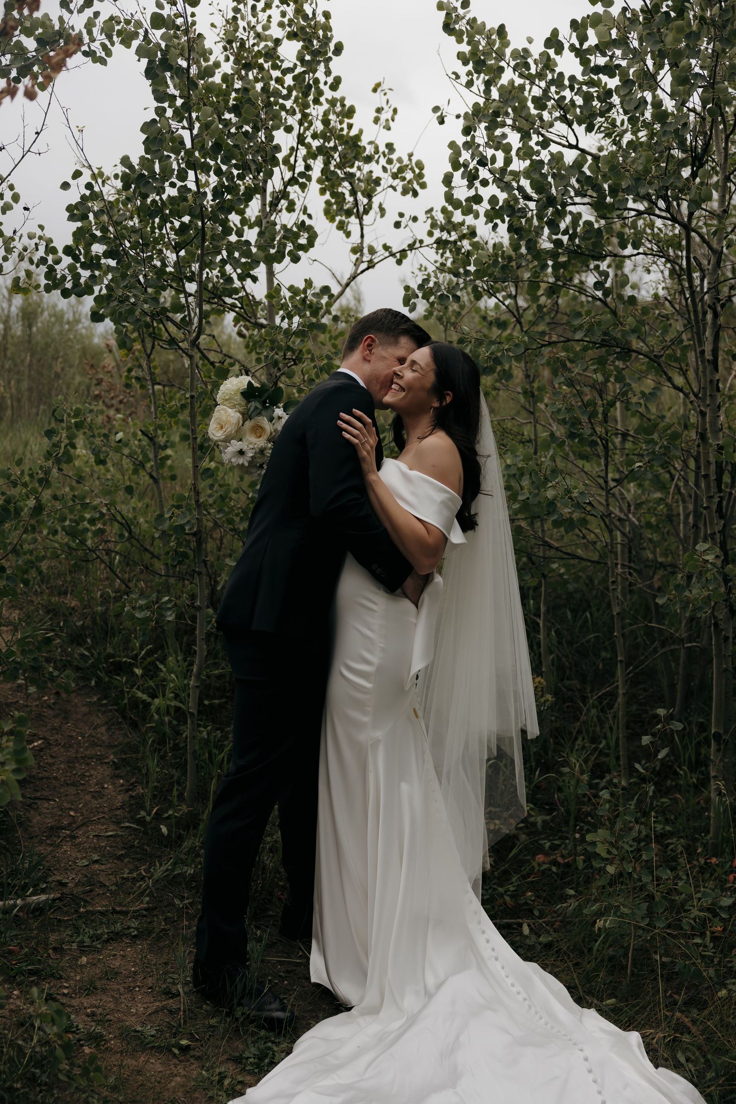 Bride and groom share a romantic embrace among lush green trees, bride wearing elegant white gown with veil.