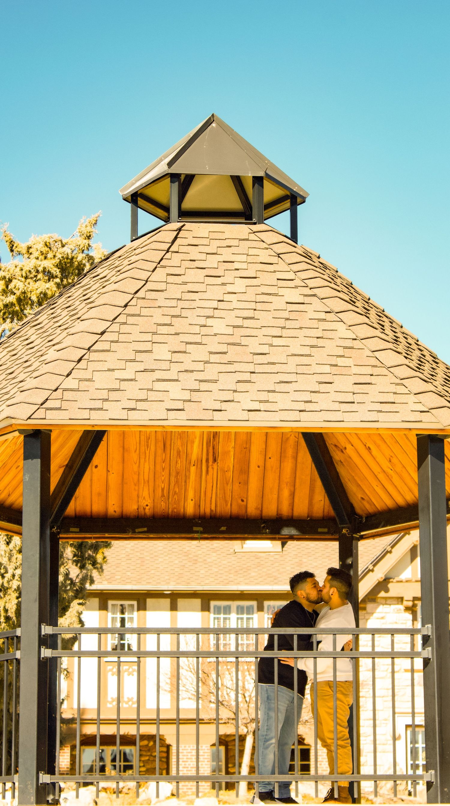 Architectural view of gazebo with wooden beams and shingle roof against clear blue sky.
