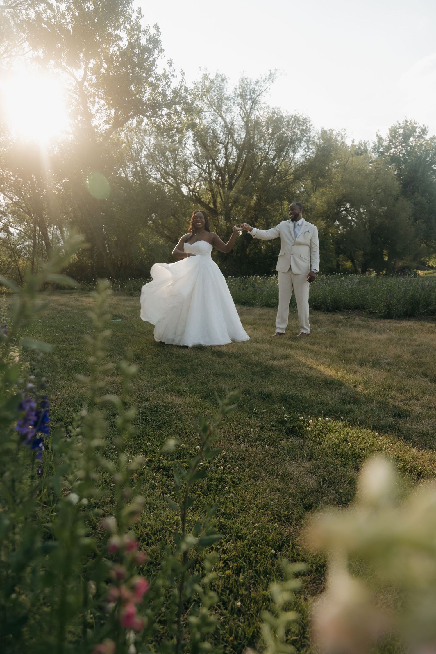 Newlywed couple dancing in a sunlit garden with wildflowers in the foreground and lush green trees behind.