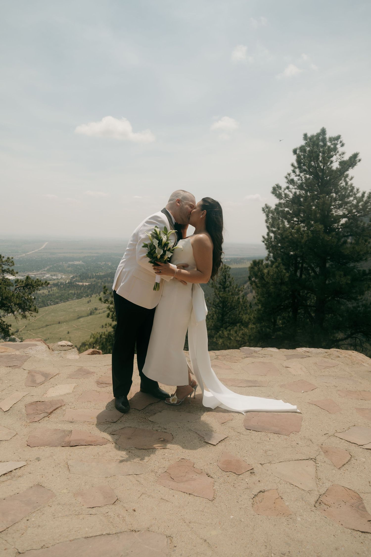 Bride and groom share a kiss on a rocky mountain overlook, bride holding a white bouquet in a flowing wedding gown.