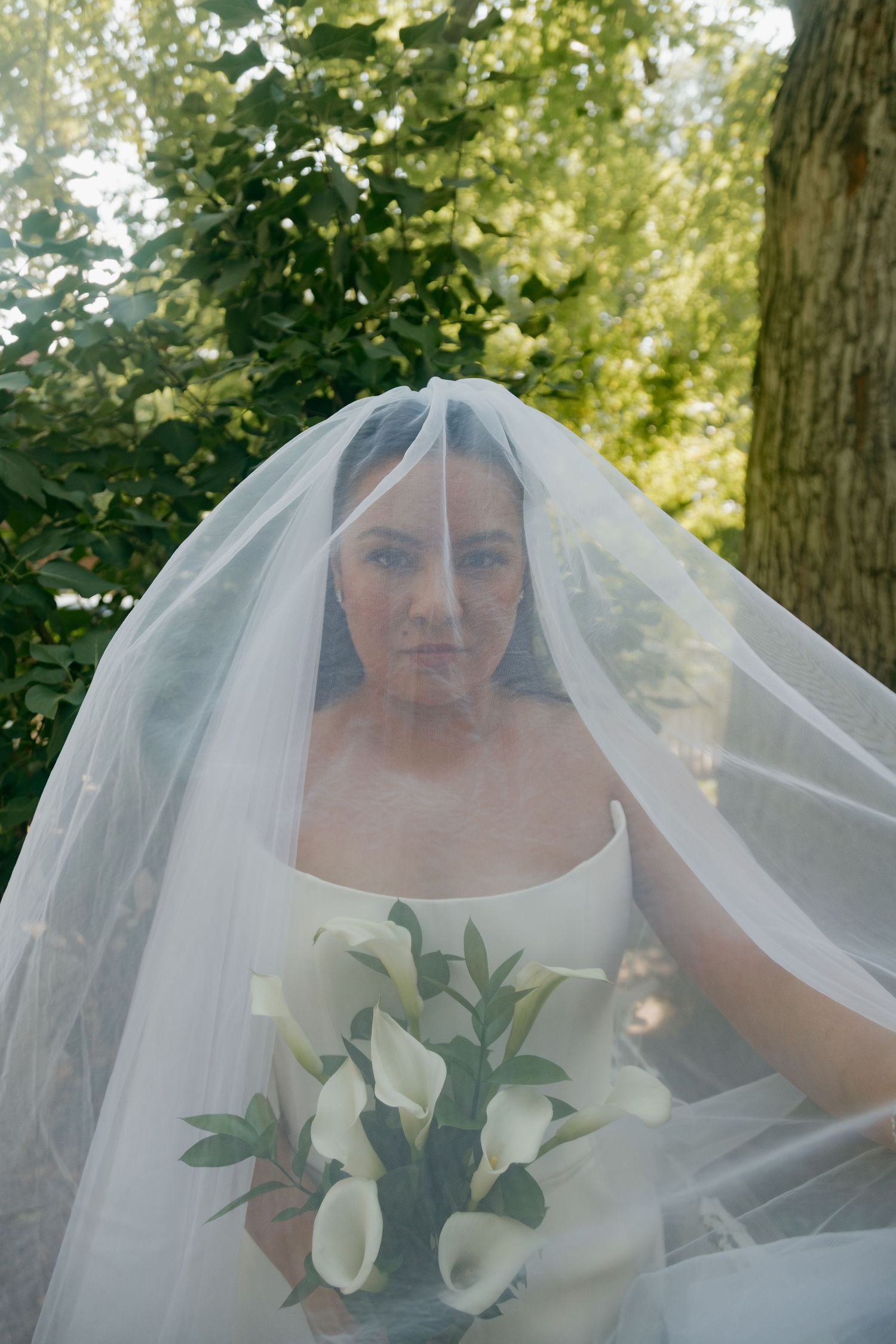 Bride wearing a white veil over her face holding a bouquet of white calla lilies outdoors.
