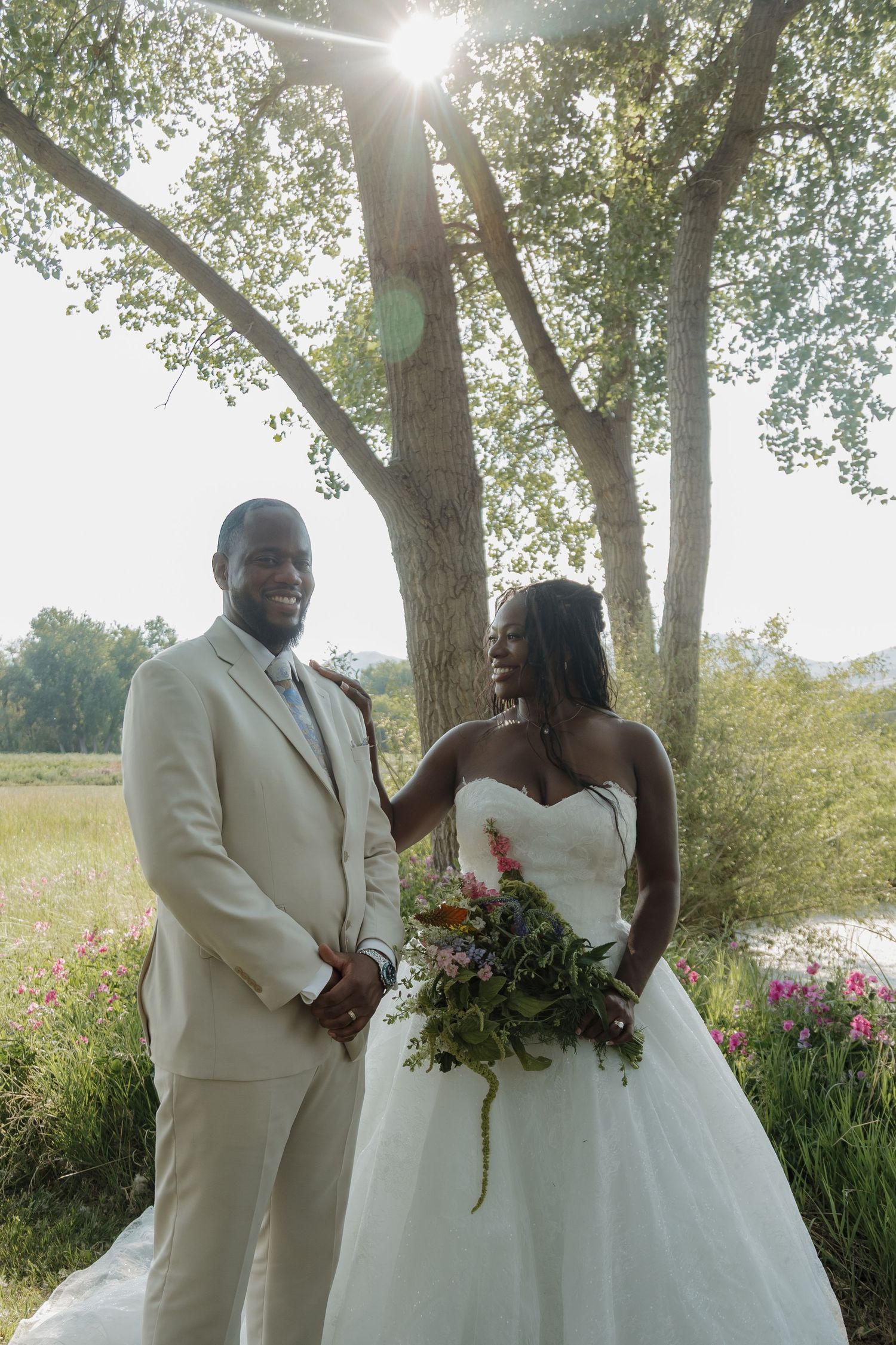 A bride and groom pose outdoors under a sunlit tree, she holds a lush bouquet in a white ballgown.