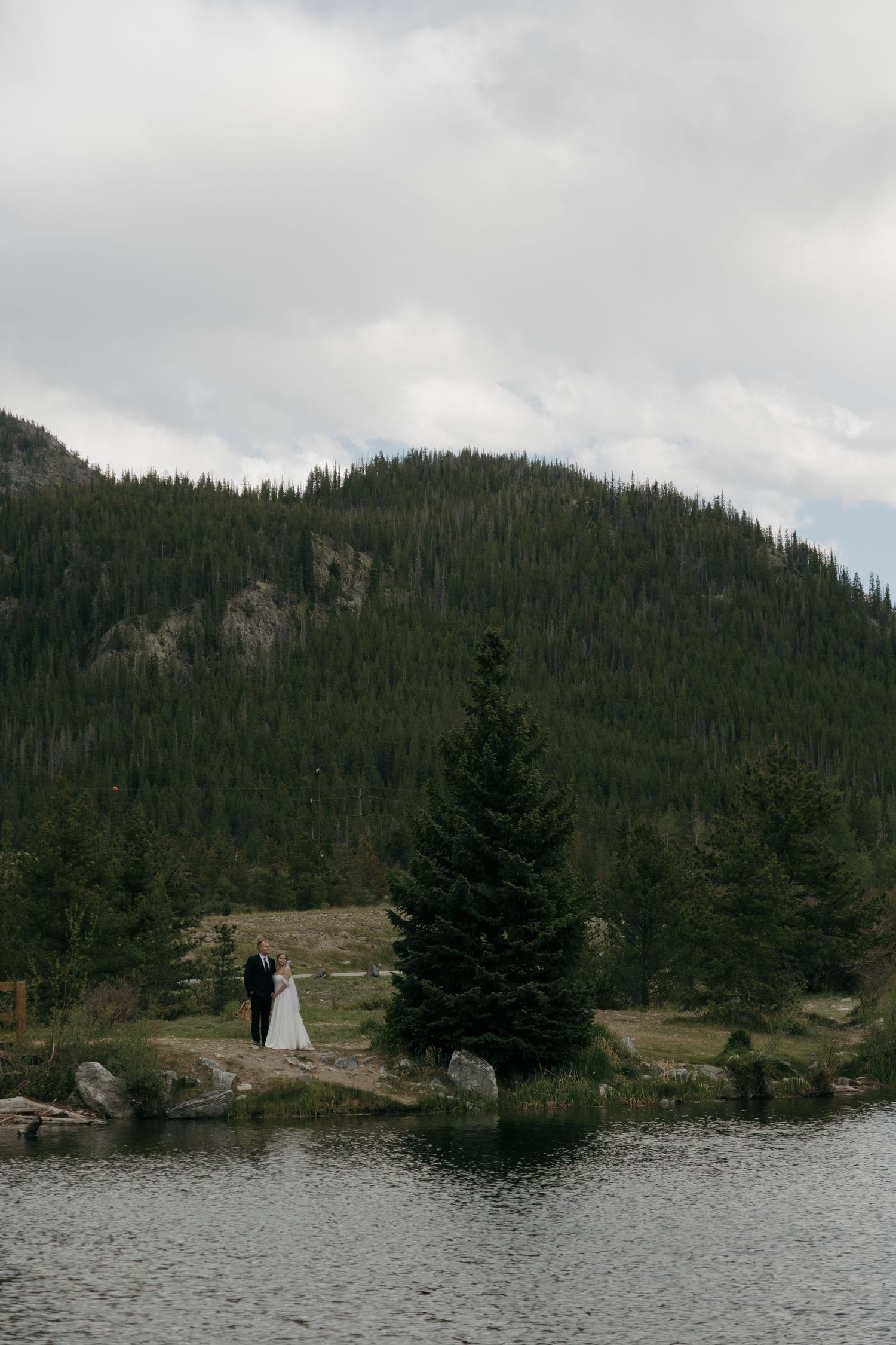 Wedding couple standing by a serene mountain lake surrounded by pine forests under cloudy skies.