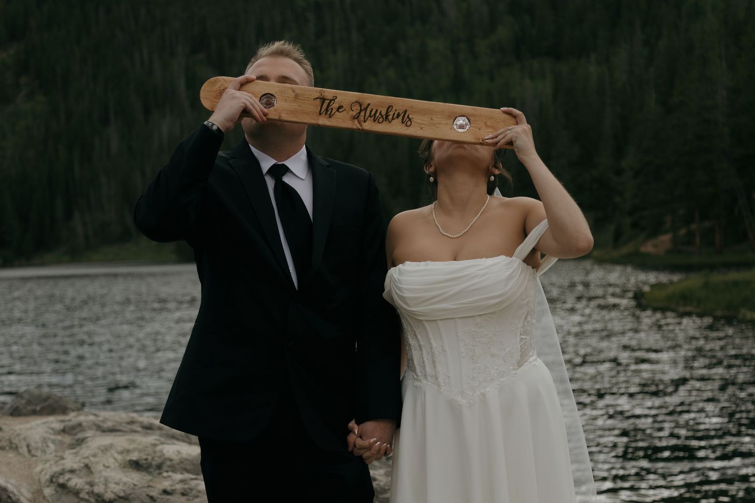 Bride and groom hold a personalized wooden sign over their faces near a mountain lake on their wedding day.