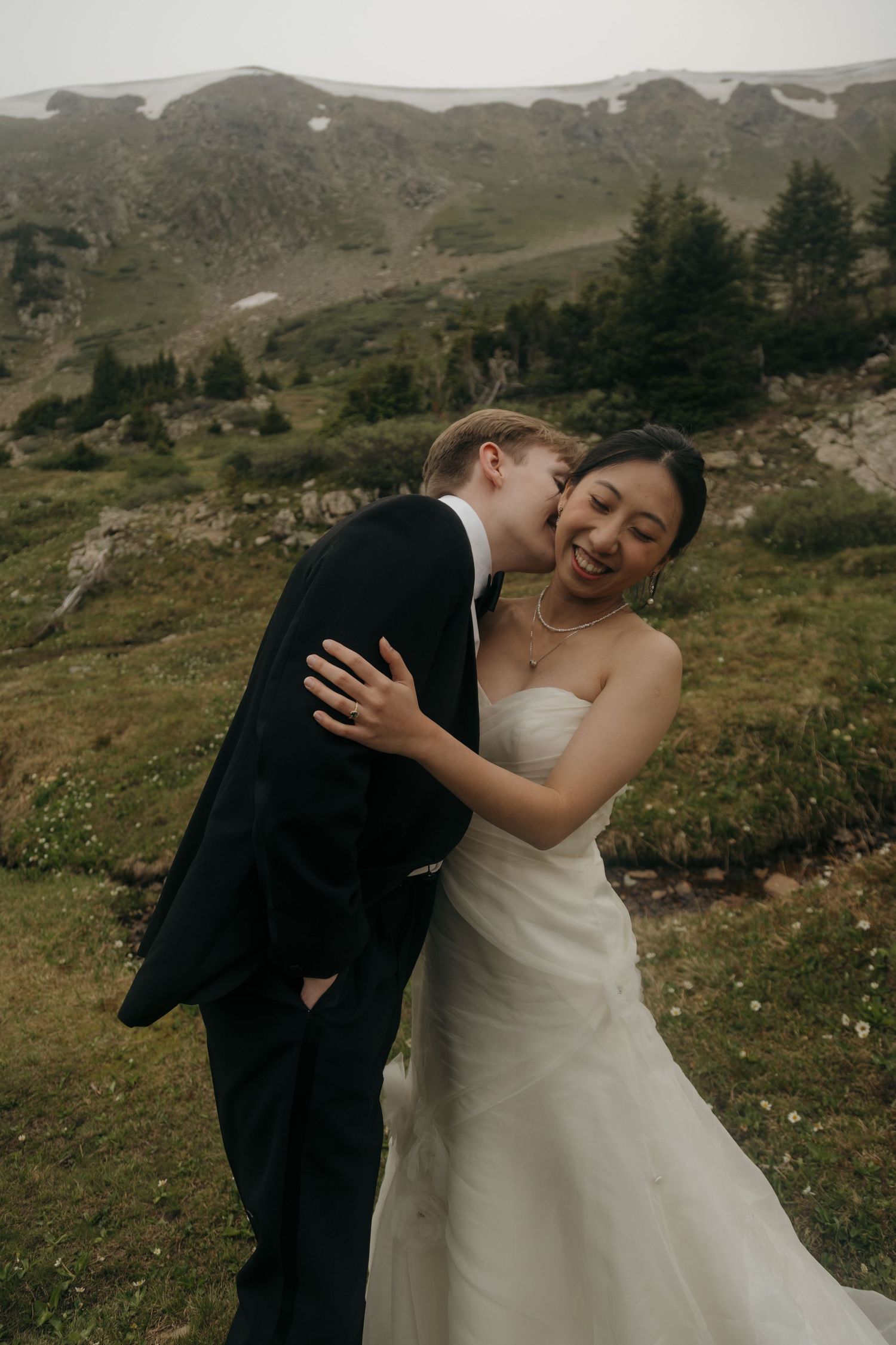 Groom kisses bride on the cheek as she laughs joyfully against a lush alpine meadow backdrop.