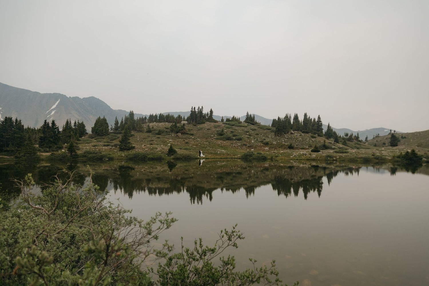 Distant bride in white gown standing beside a serene alpine lake reflecting misty mountain scenery.