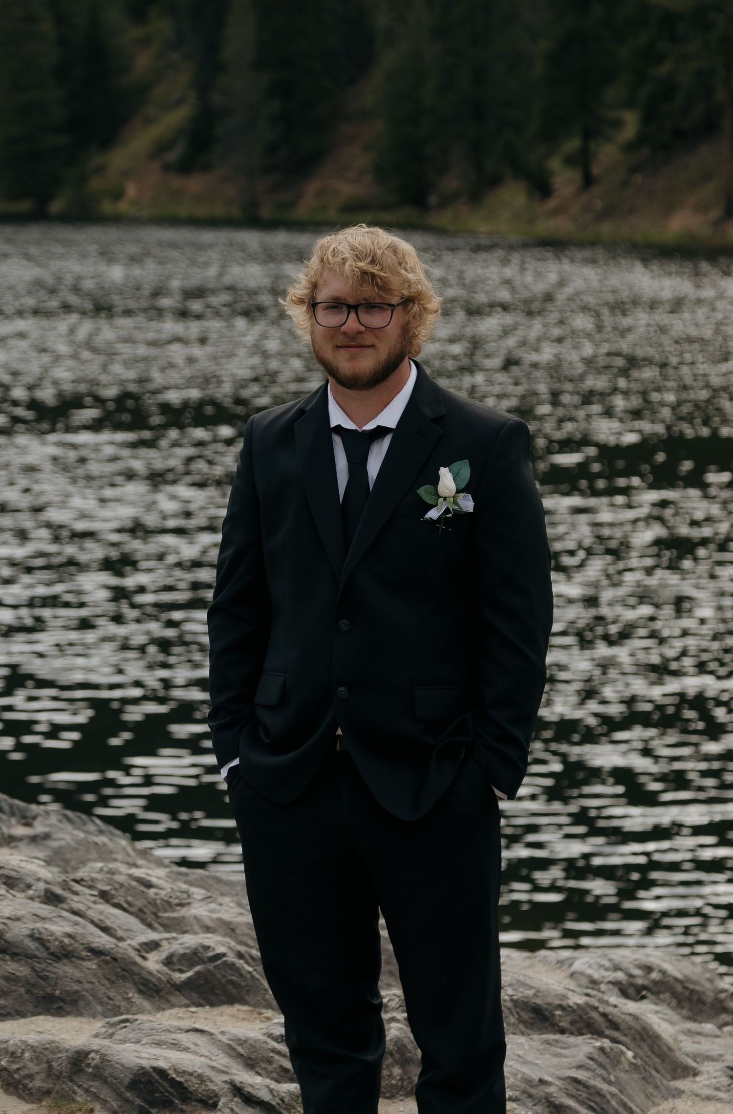 Groom in black suit with boutonniere stands on rocky shore by mountain lake.