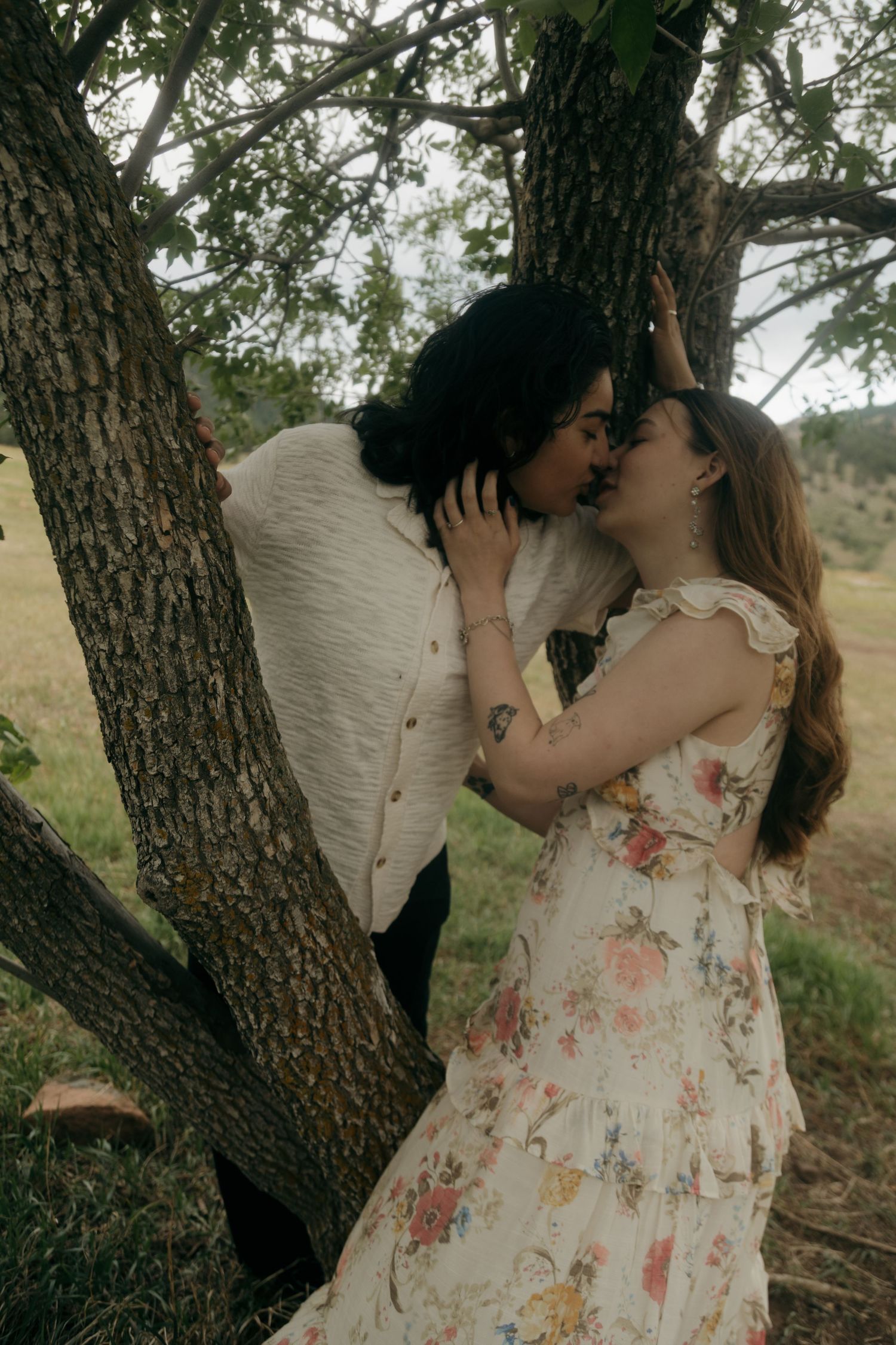 A couple shares a romantic kiss leaning against a tree trunk in a natural outdoor setting.