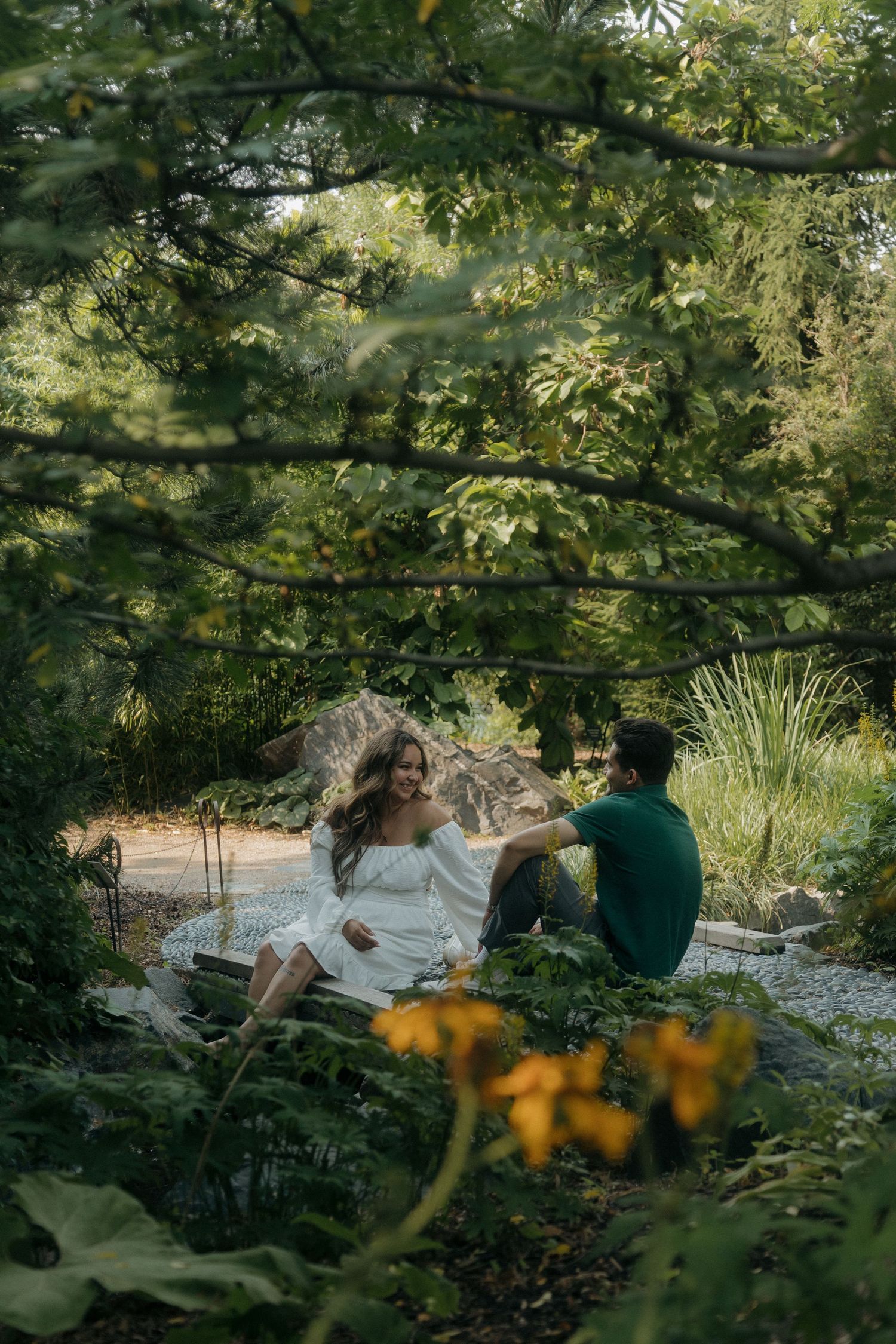 Couple relaxing together in a lush garden, woman in white dress and man in green shirt sitting on the ground.