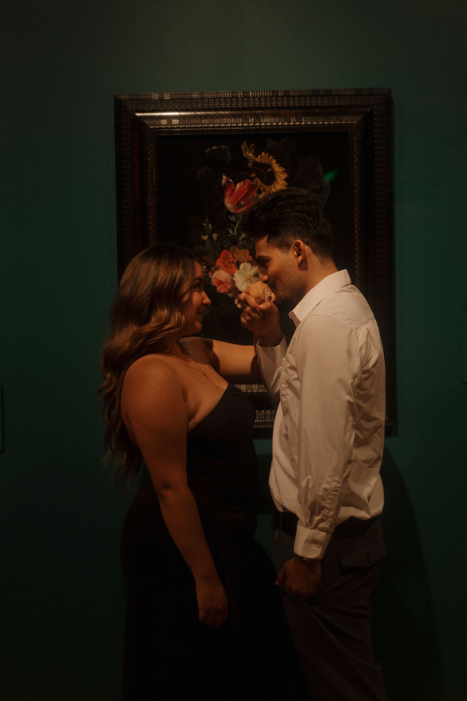 Couple sharing an intimate moment in front of a dark floral still life painting in a moody gallery.