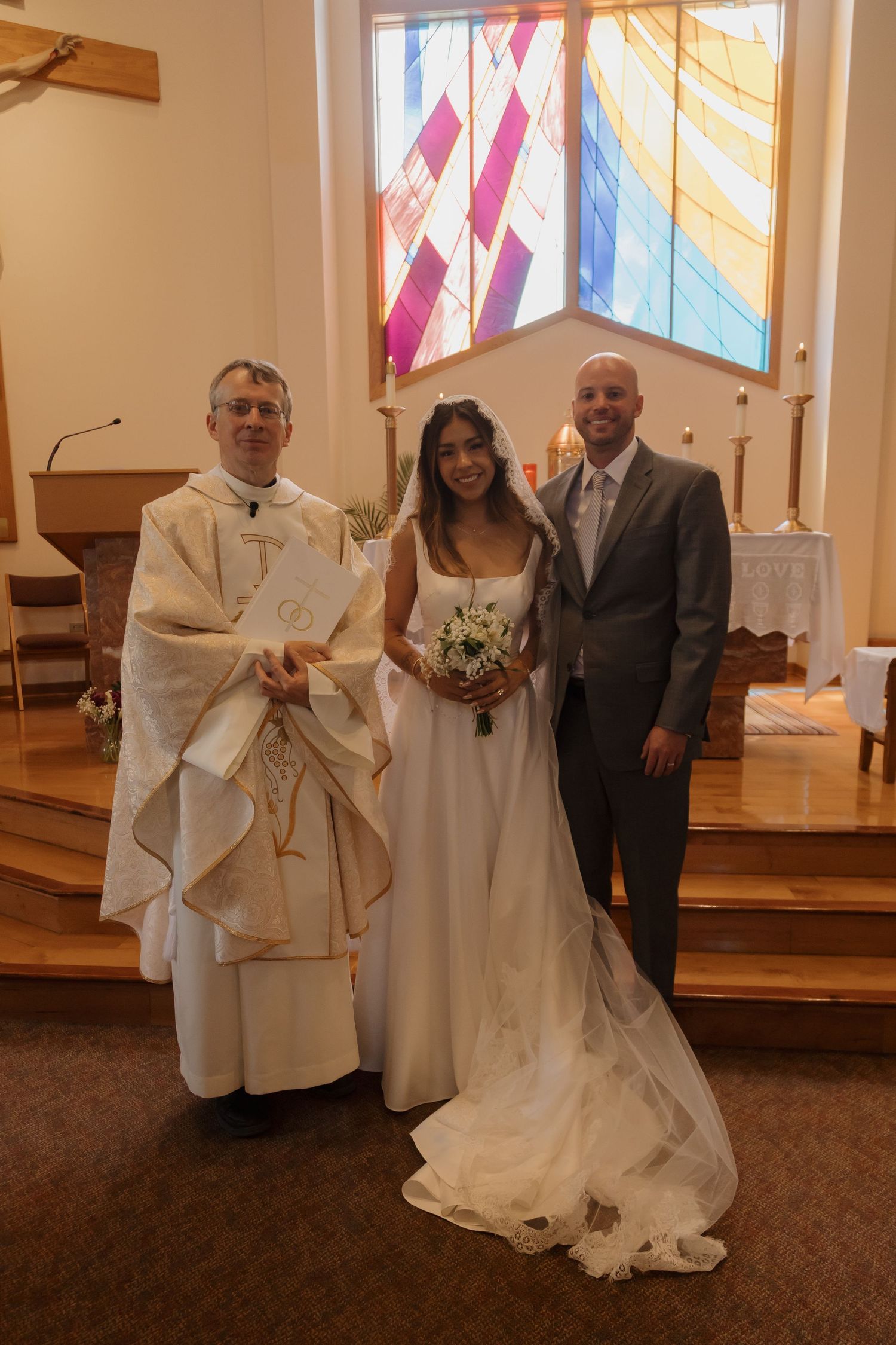 Newlywed couple posing with priest in front of colorful stained glass window after Catholic wedding ceremony.