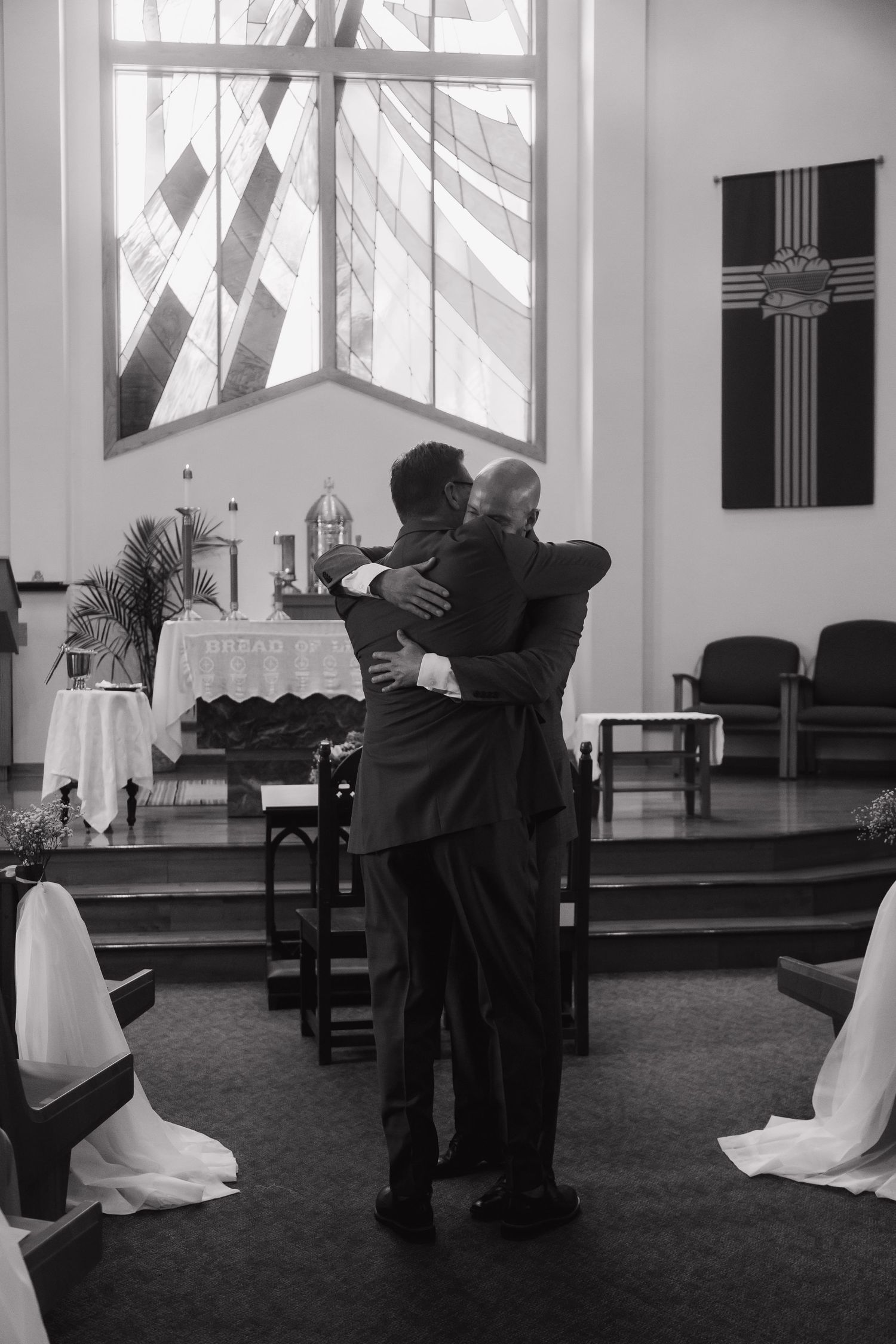 Two men in suits share an emotional hug at a church altar during a wedding ceremony.