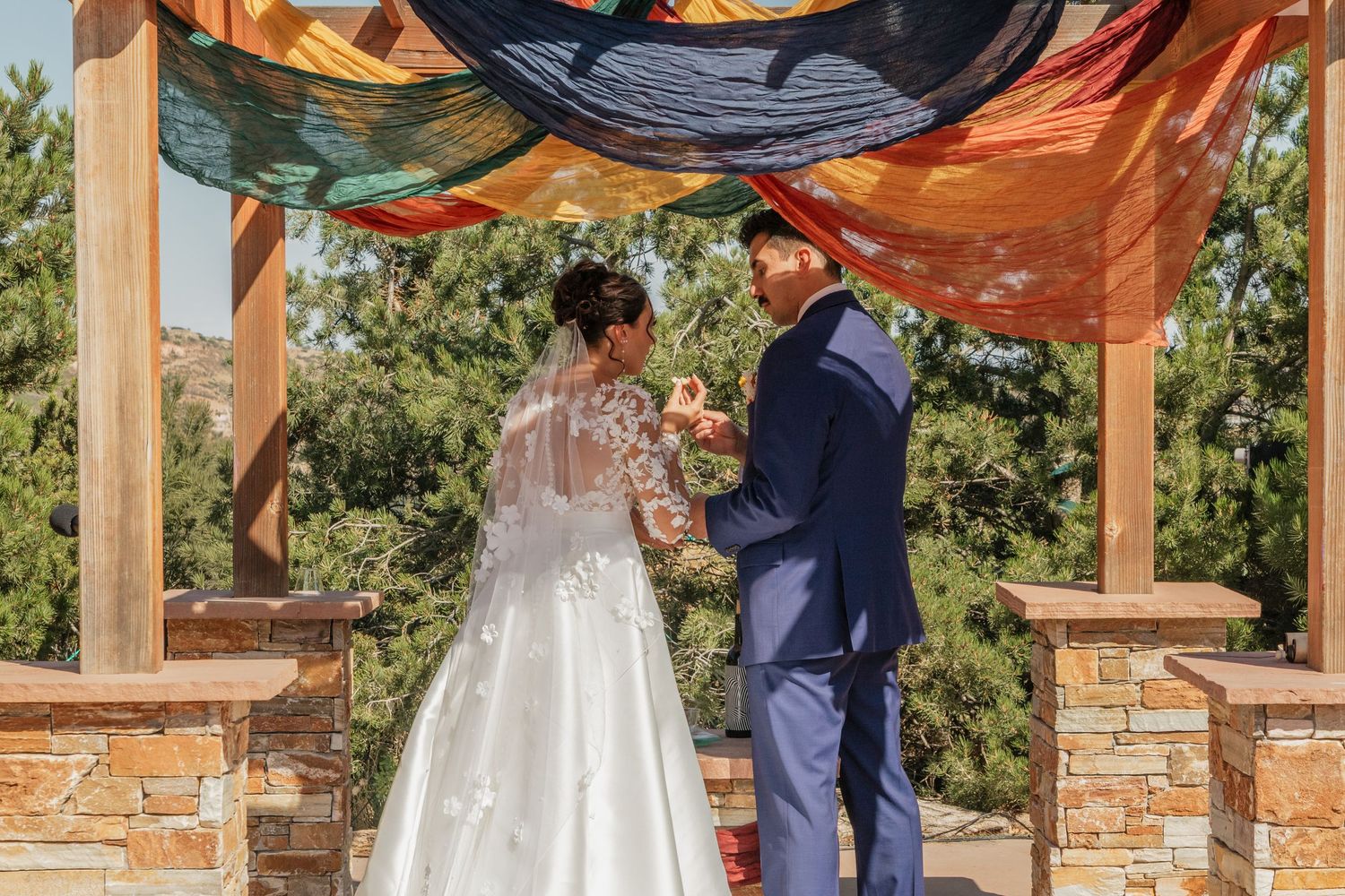 Wedding ceremony setup with colorful fabric draped over wooden beams against a natural outdoor backdrop.