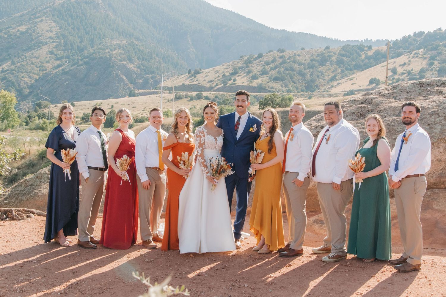 Group photo of wedding party in earth-toned attire with mountains in background.