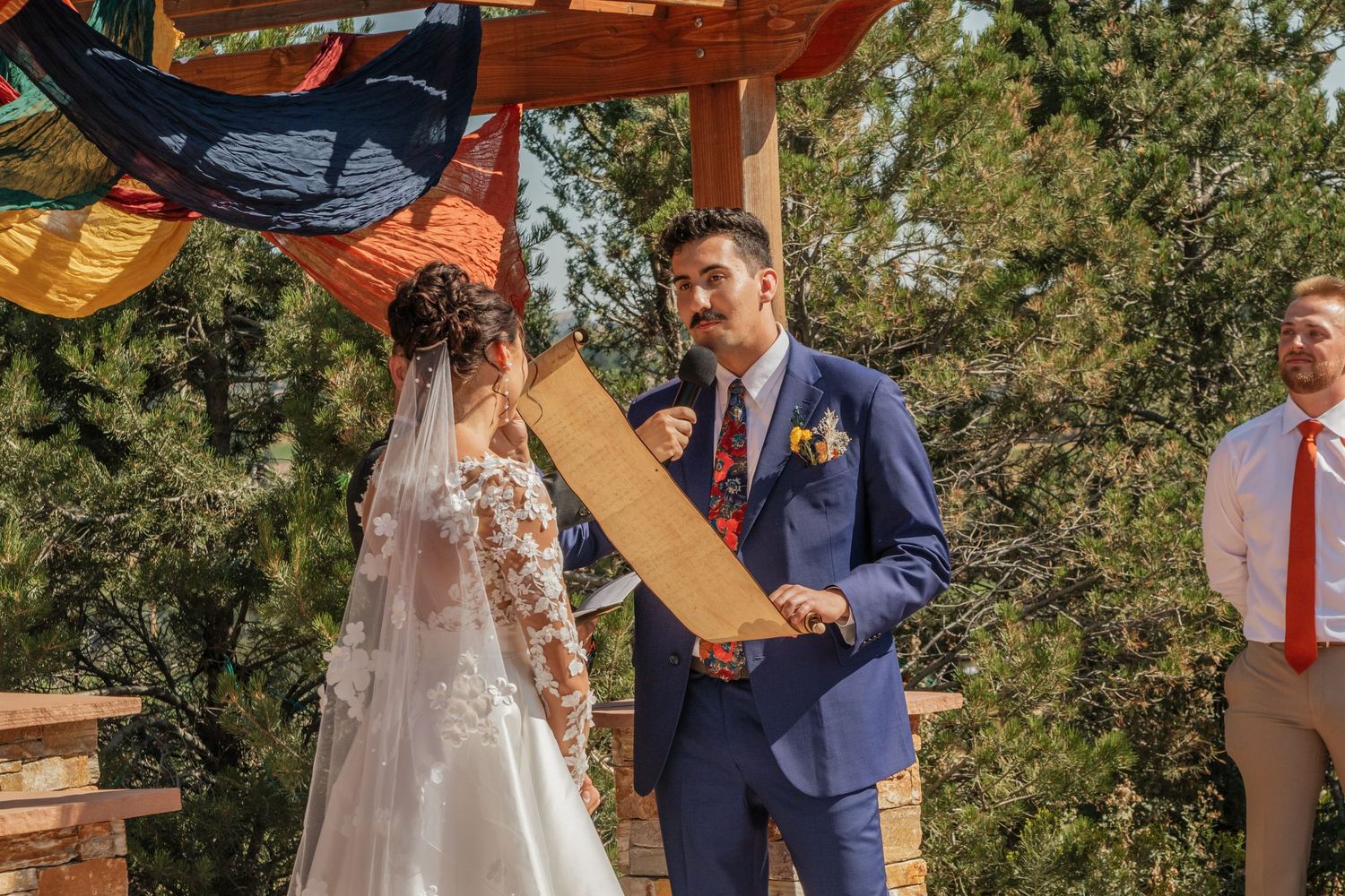 Wedding ceremony under a wooden pergola with colorful fabric draping and natural greenery backdrop.
