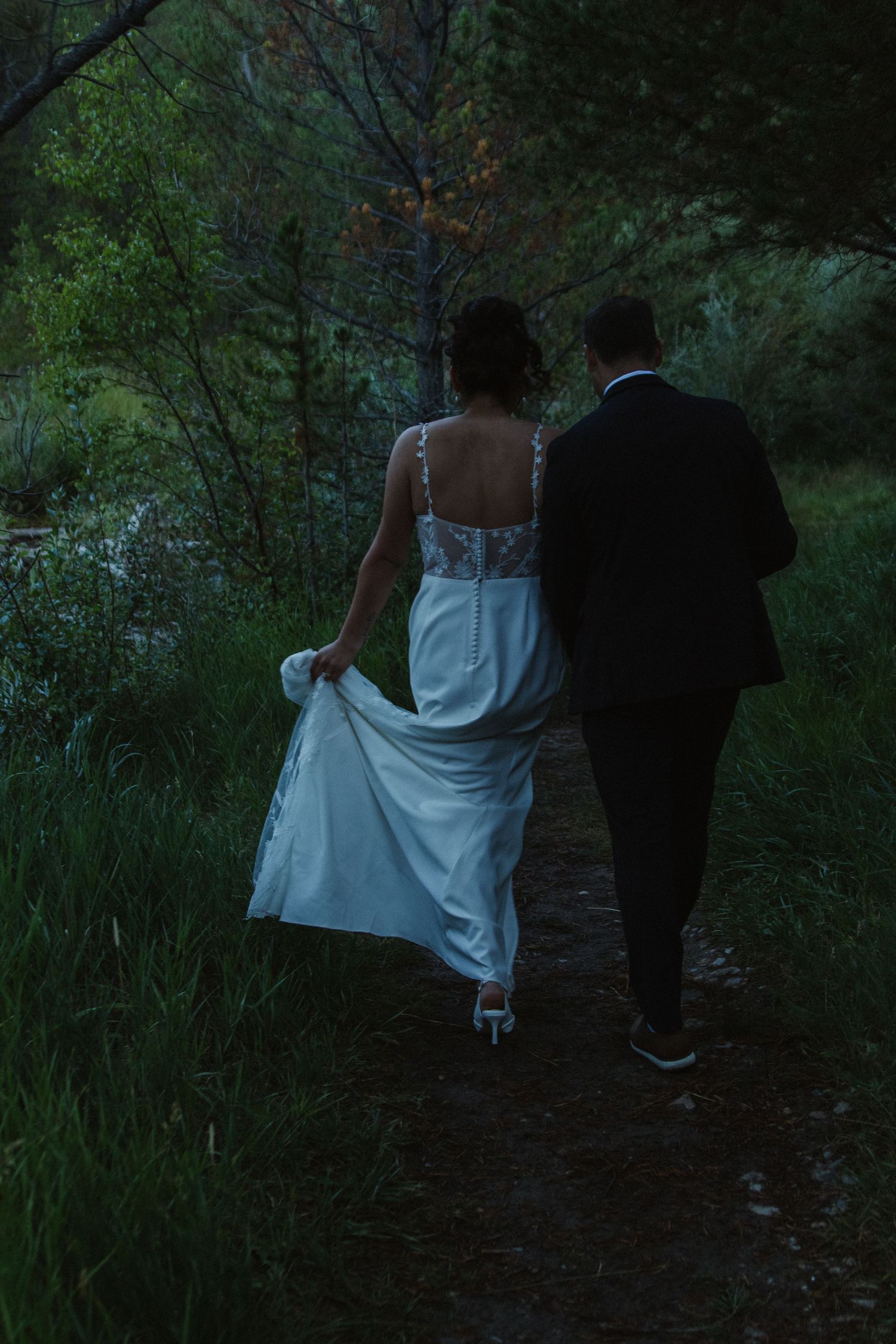 A couple walks along a dark wooded path in wedding attire during dusk.