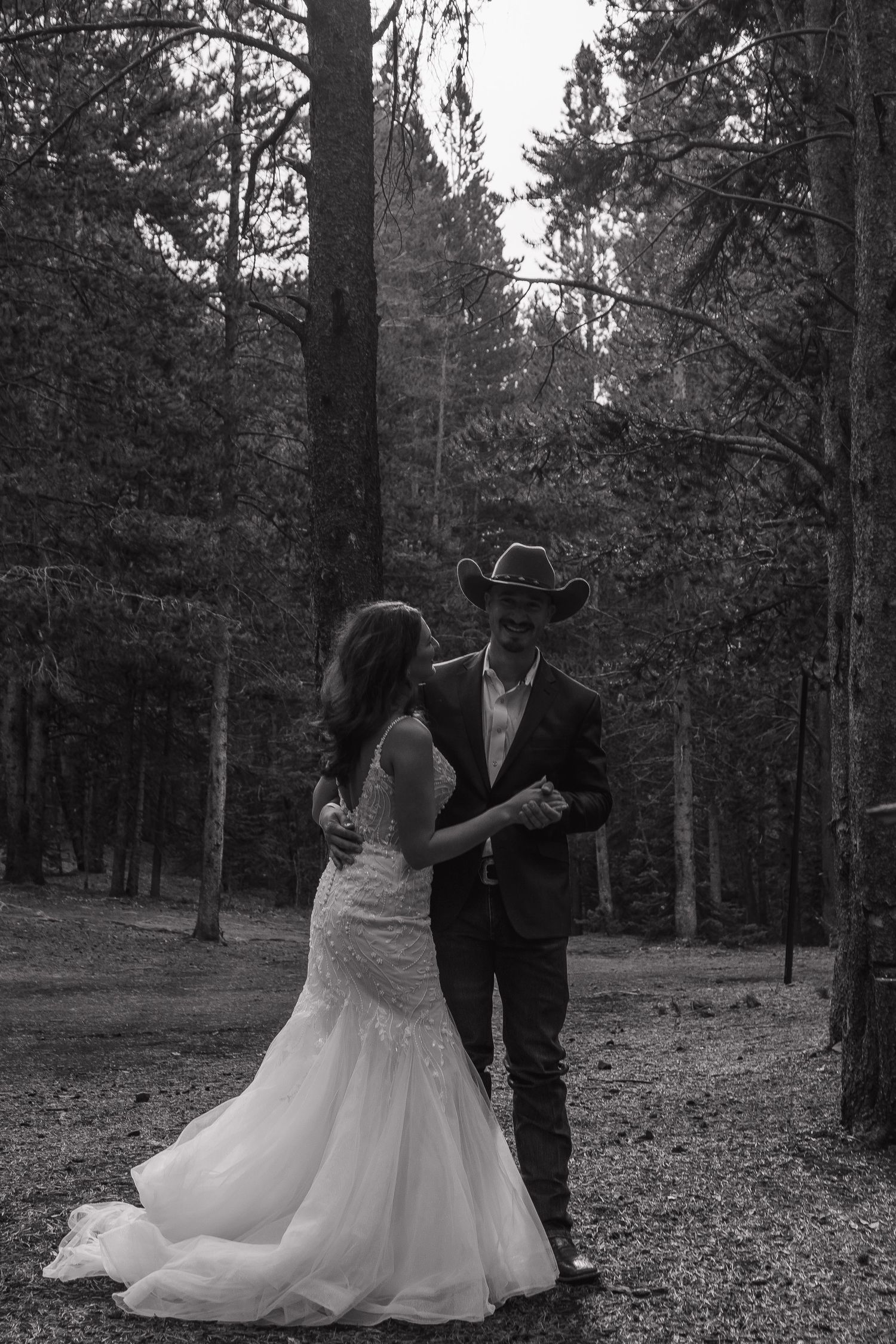 A romantic black and white wedding photo of a couple dancing among tall pine trees in a forest setting.