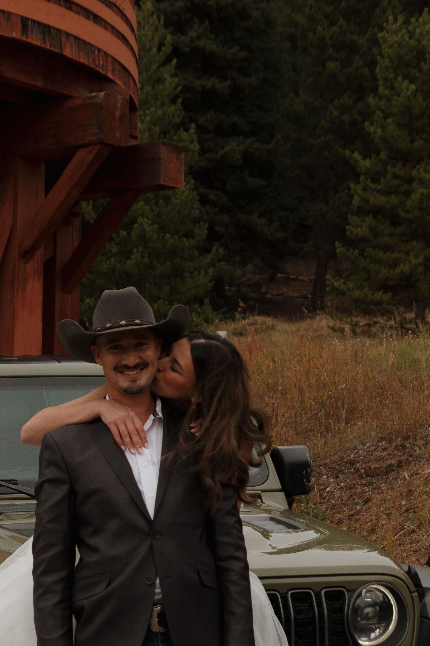 A couple shares an intimate moment near a vintage Jeep with wooden structures in the background.