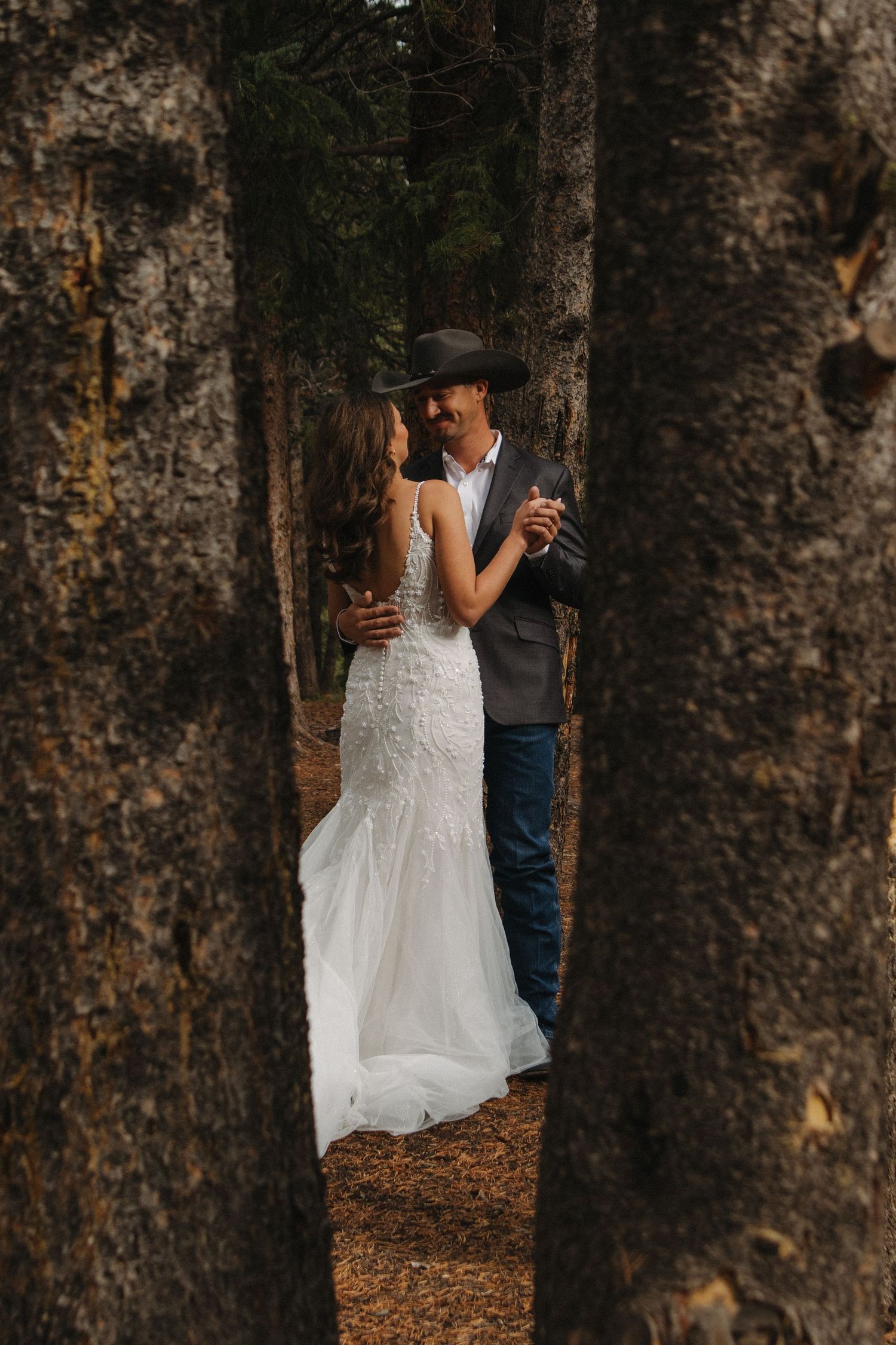 A romantic wedding moment captured between trees with a white wedding dress and western attire.