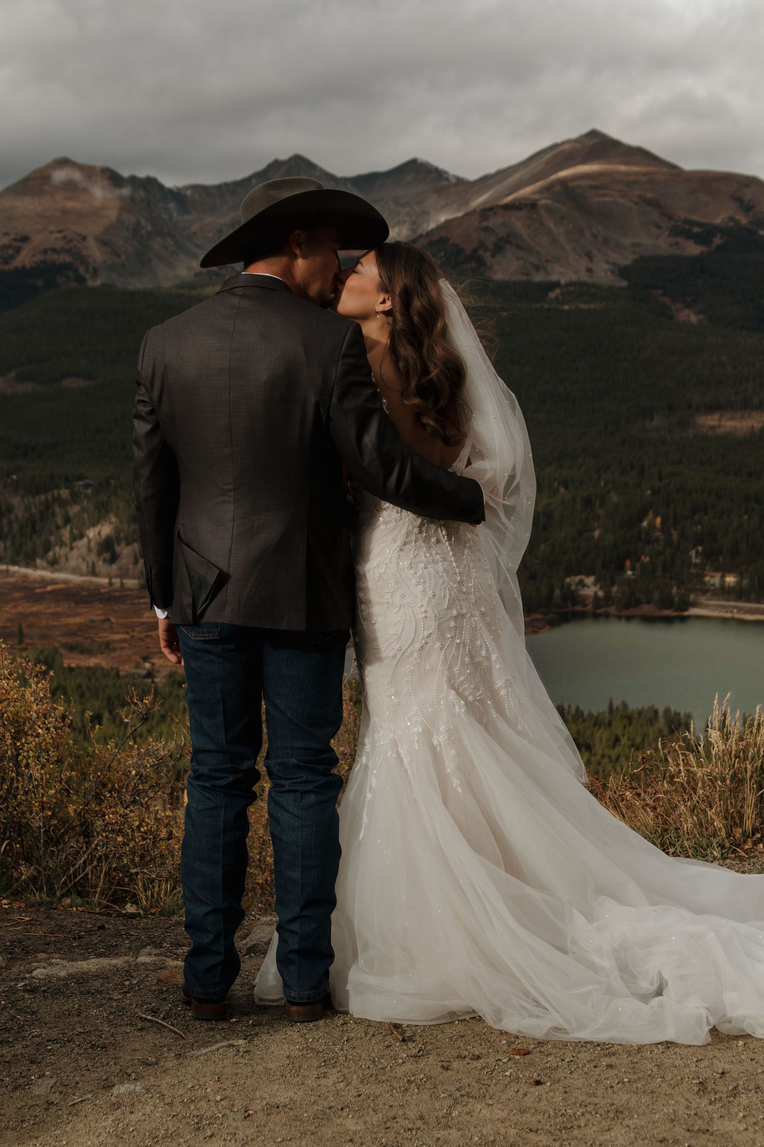 A couple in wedding attire shares an intimate moment overlooking a scenic mountain lake landscape.
