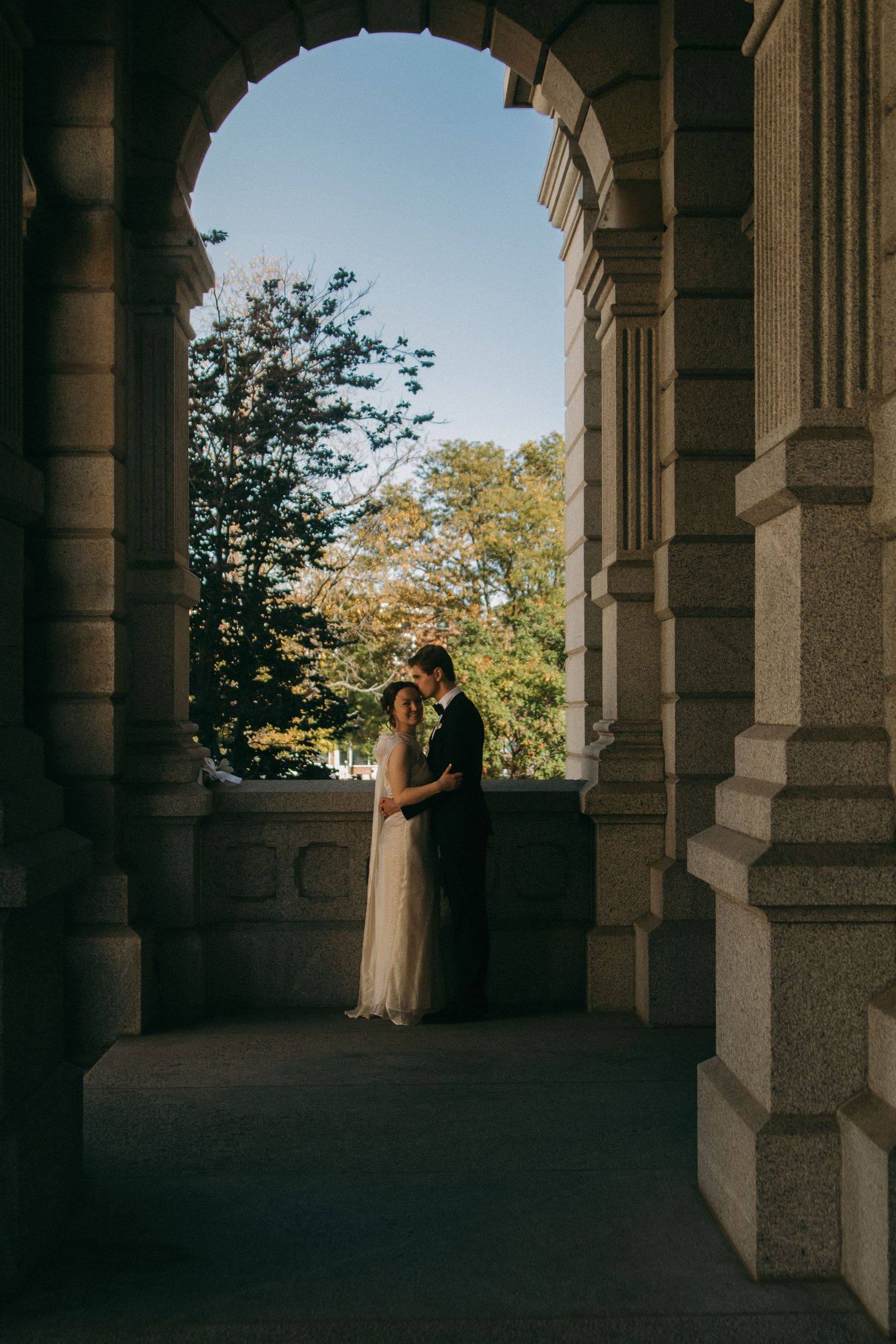 Wedding couple silhouetted under stone archways with trees in background.