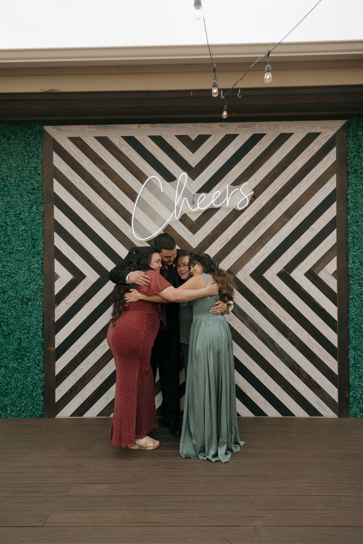 Four wedding guests share a group hug in front of geometric black and white Cheers neon sign backdrop.