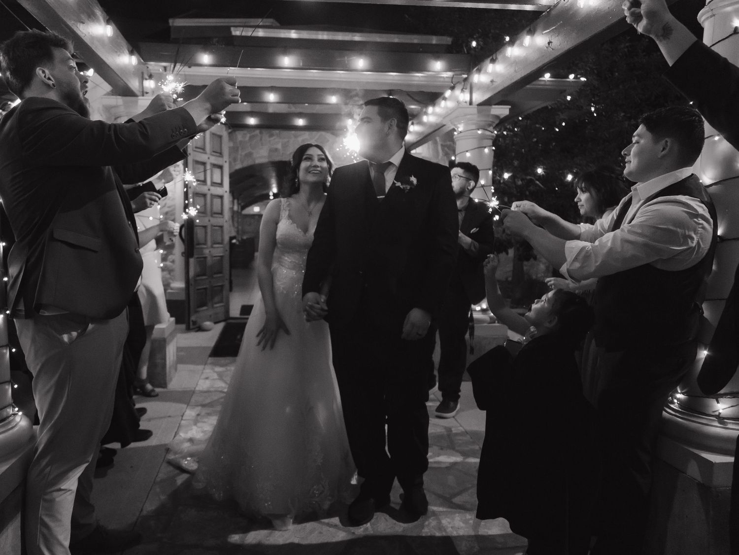 Black and white photo of smiling newlyweds walking through a sparkler tunnel at their reception.